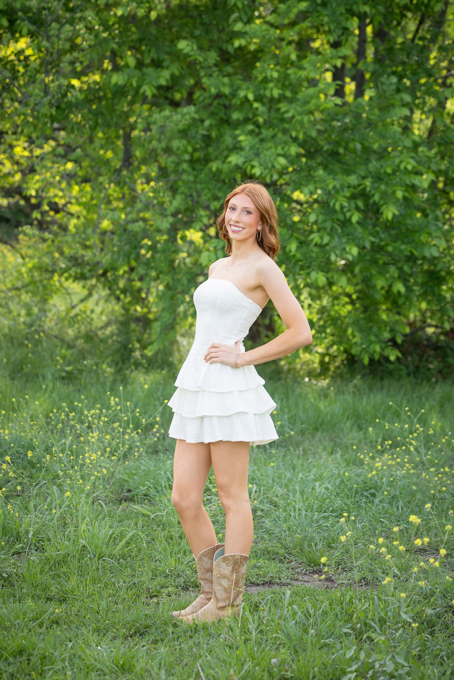Young woman in white strapless tiered mini dress and cowboy boots posing in a lush green meadow with wildflowers.