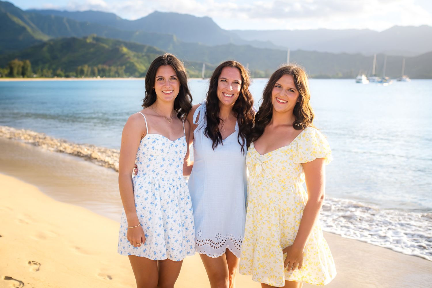 Three people in summer dresses pose together on a tropical beach with mountains and sailboats in the background.