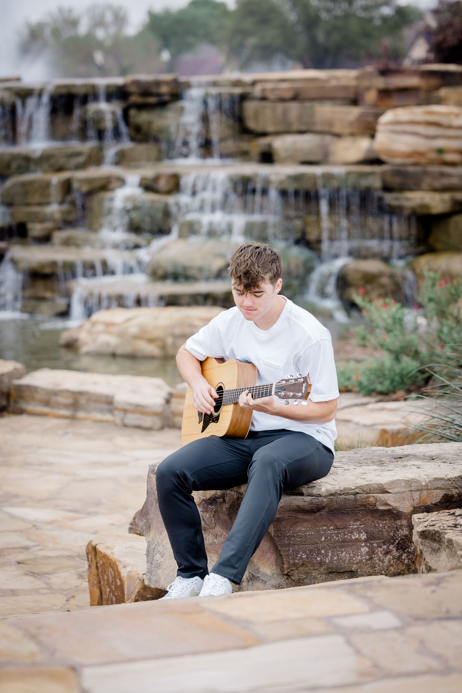 Musician playing acoustic guitar while sitting on rocks near cascading waterfall.