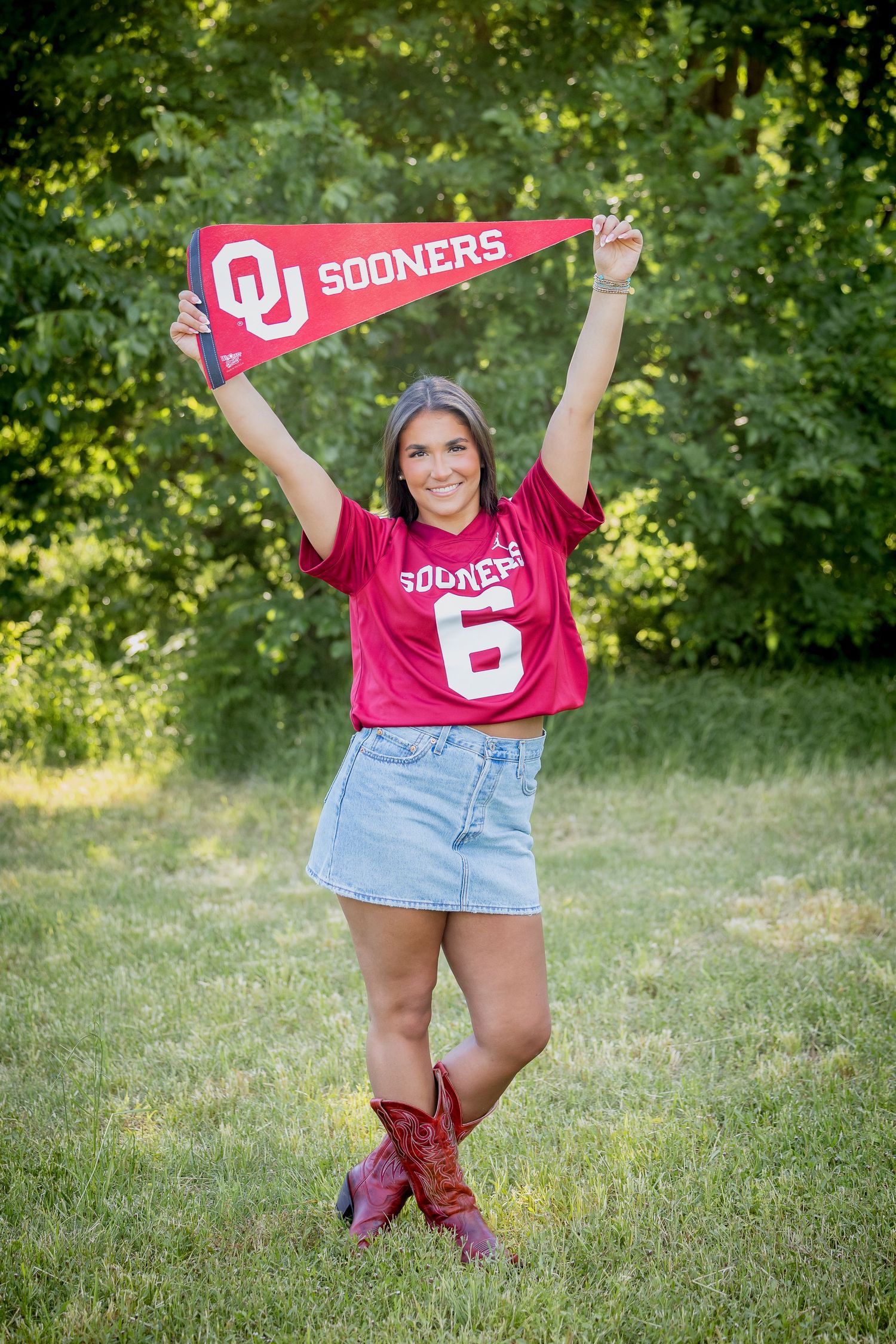 A smiling young woman holds a Sooners pennant overhead, wearing a red OU jersey, denim skirt, and cowboy boots outdoors.