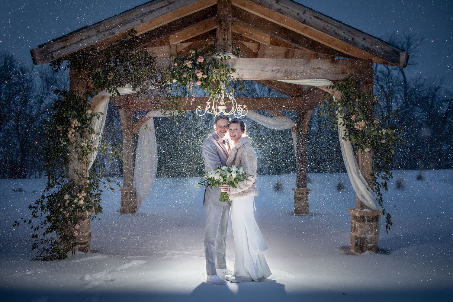 A romantic couple in wedding attire poses together under an illuminated rustic wooden pergola at twilight.