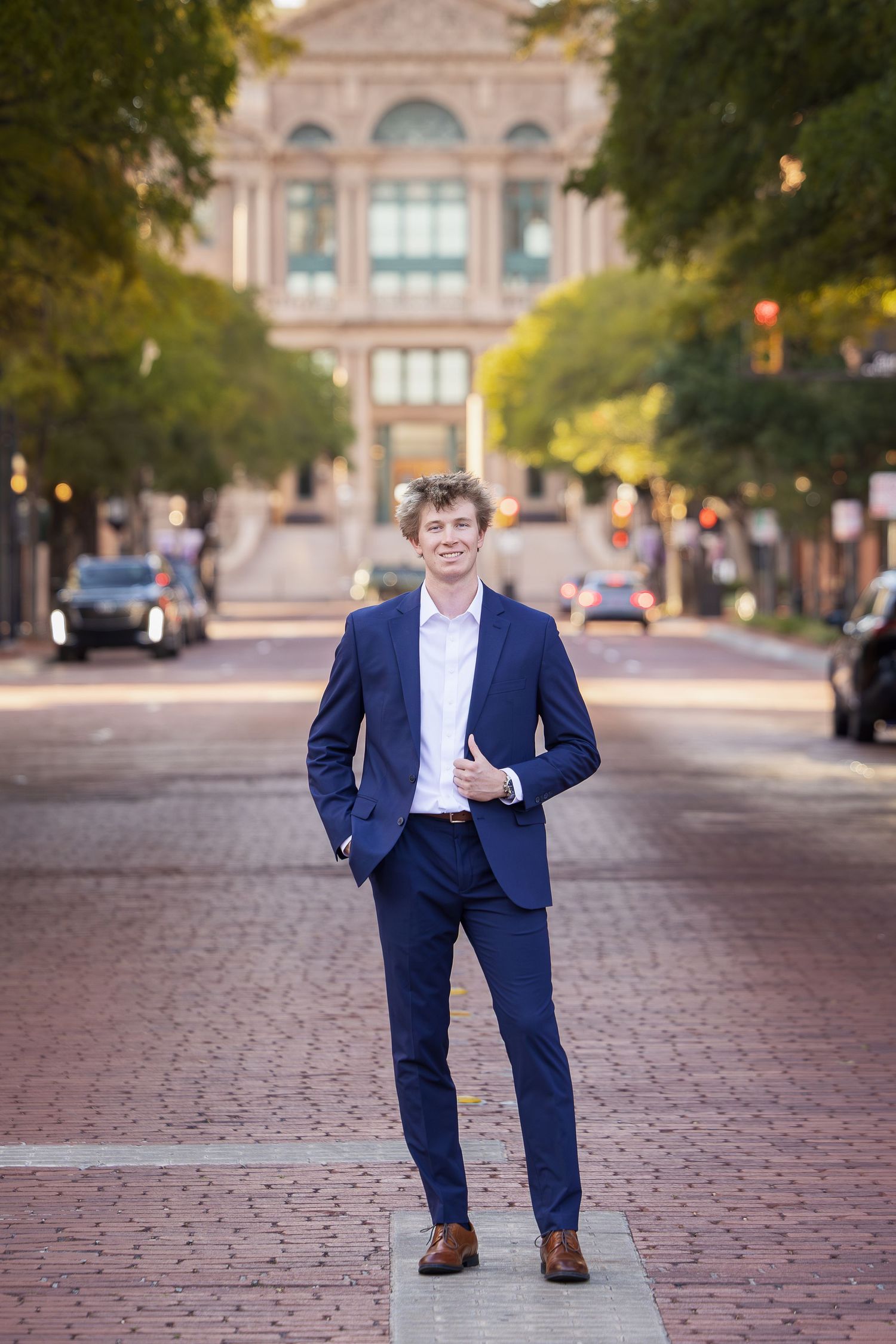 Young man in navy blue suit standing confidently on brick plaza with grand building and tree-lined street in background.