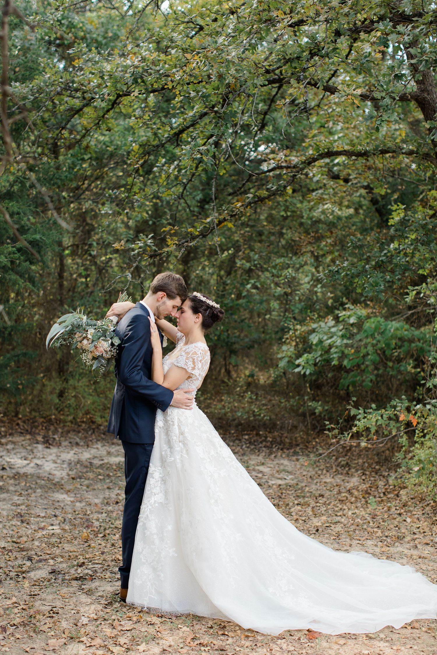 A couple in wedding attire share an intimate moment on a tree-lined dirt path in a wooded setting.
