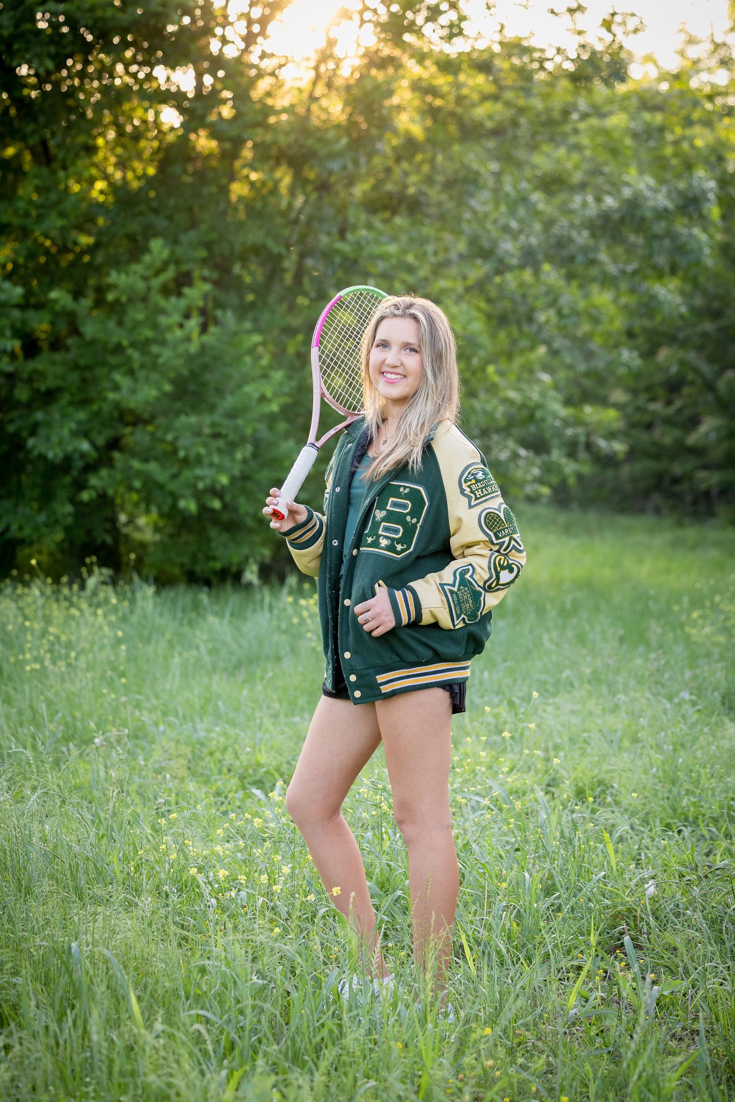 Smiling blonde teen in green varsity jacket holds tennis racket in sunlit green field for senior portrait.