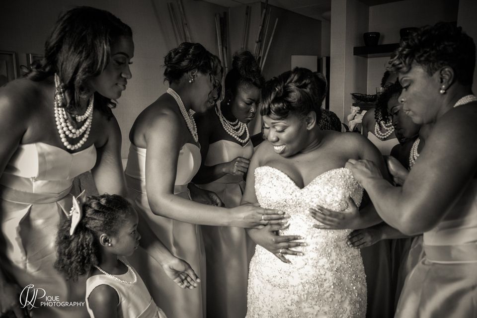 A candid black and white photo captures bridesmaids helping with wedding dress preparations.
