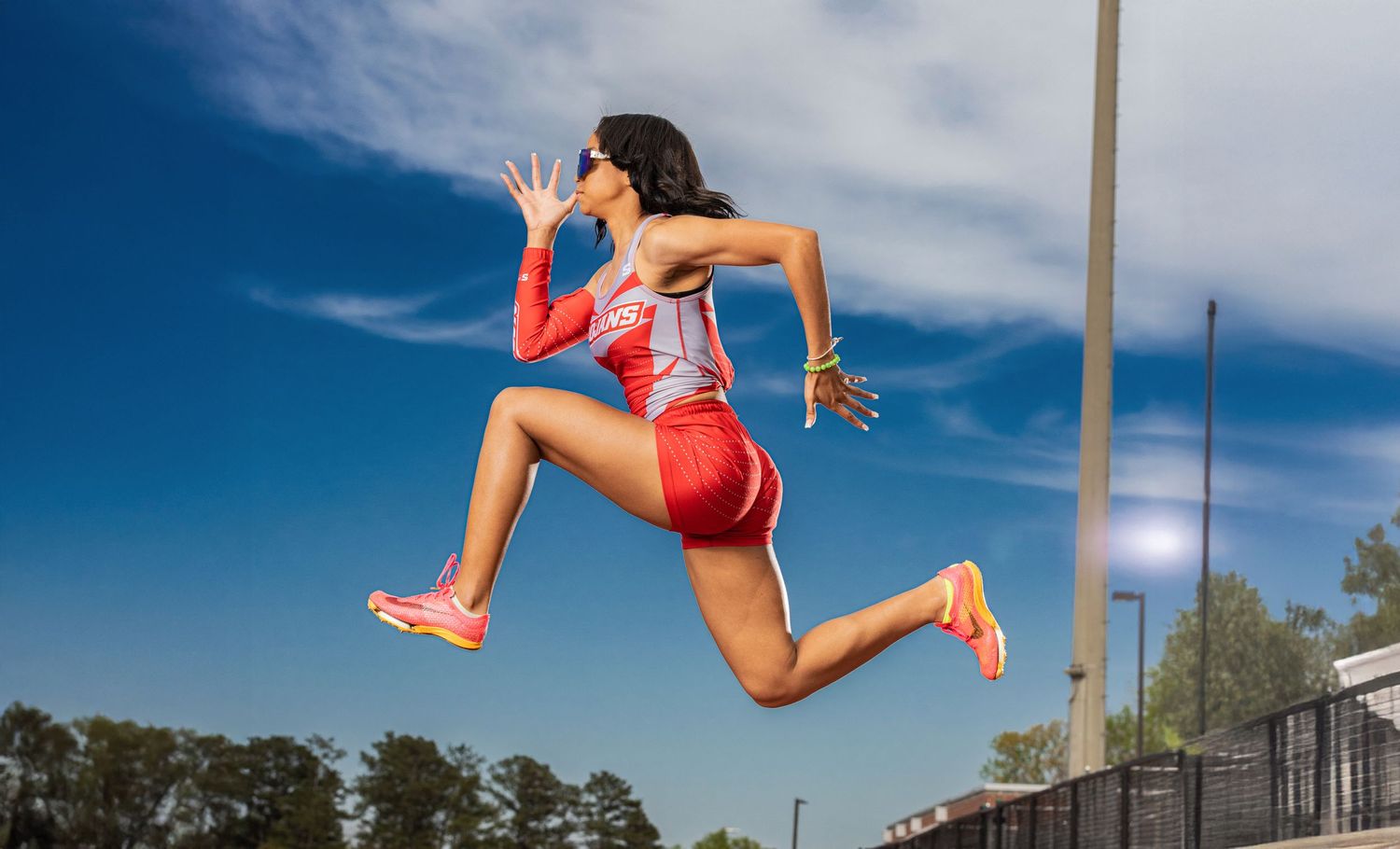 Athletic figure leaping high against a blue sky in red athletic wear during an outdoor track and field event.