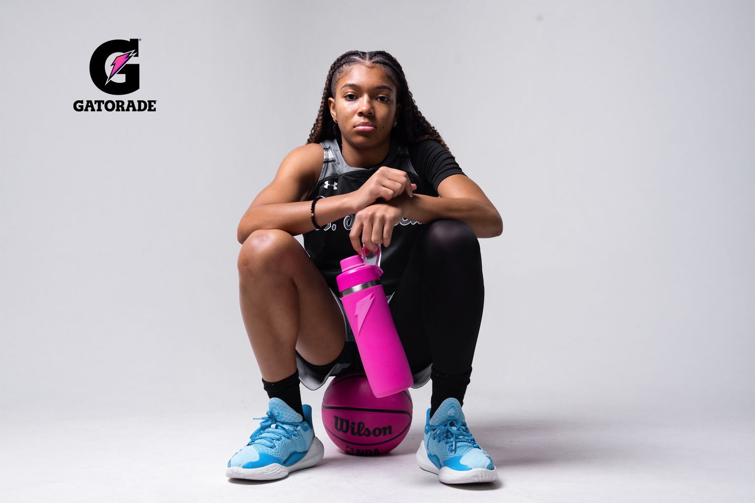 Athlete in black outfit and blue shoes posing with pink Gatorade bottle against white background.