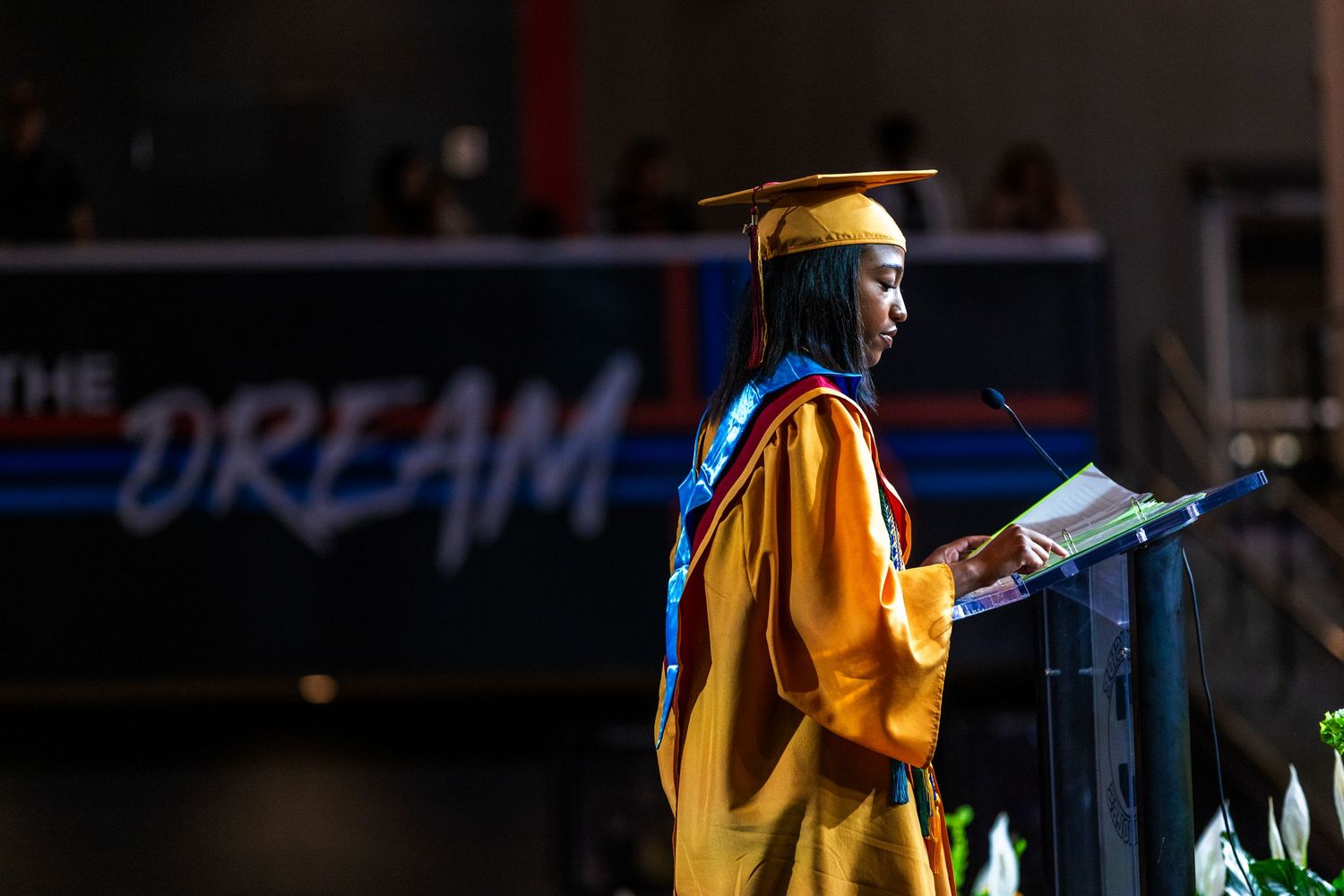 Graduate in gold and blue regalia speaking at podium during outdoor ceremony.