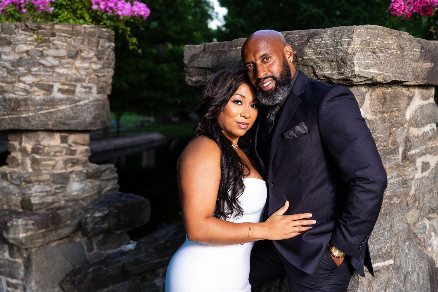 A couple poses for romantic wedding photos against a stone wall with purple flowers.