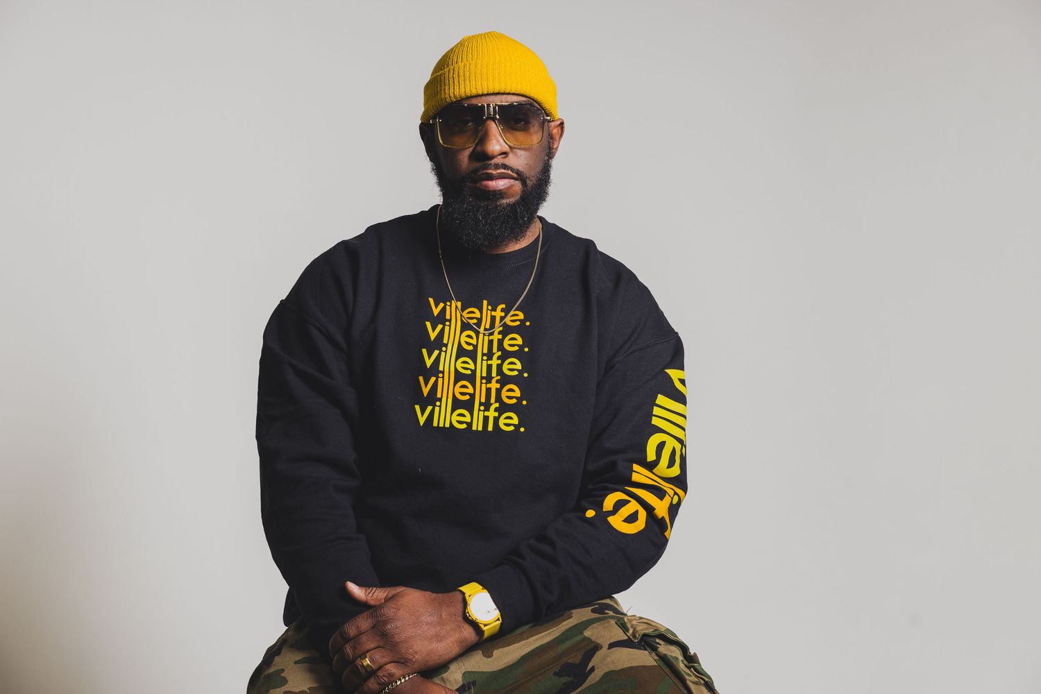 Person wearing a yellow beanie and black sweatshirt with yellow text sits against grey background.