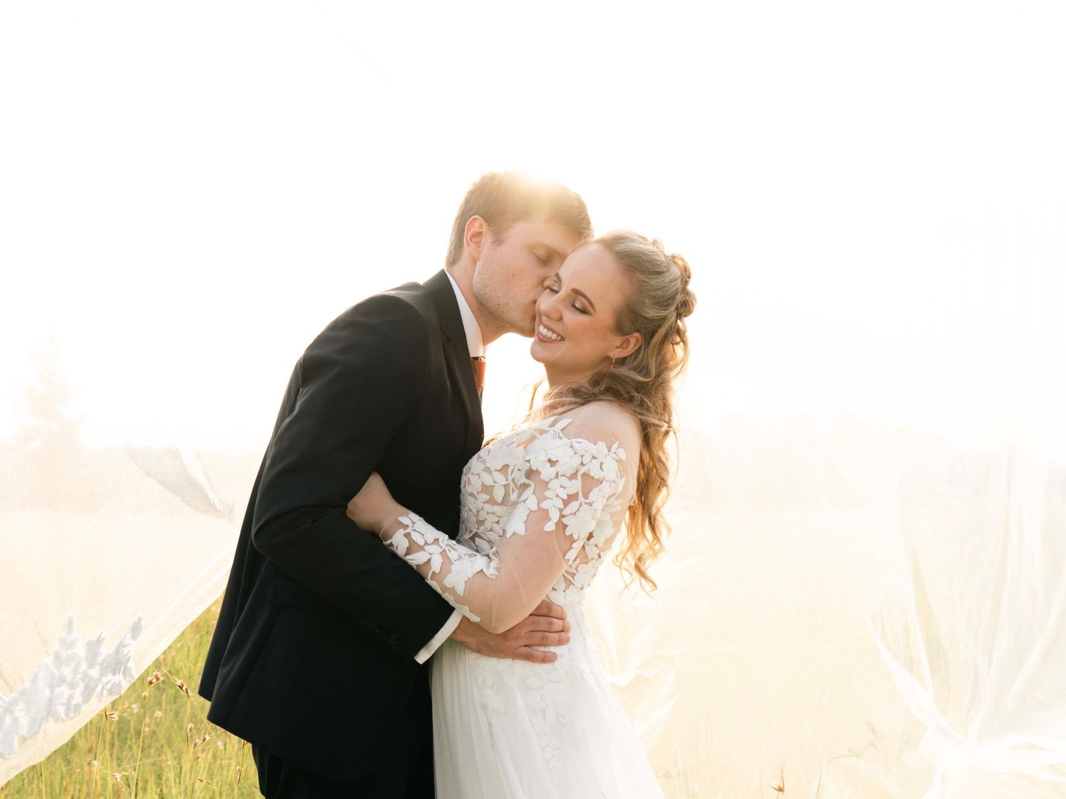 Groom kissing the bride's cheek in a sunlit field at their wedding