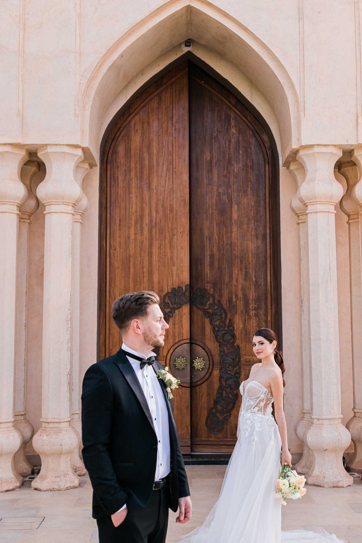 A wedding couple poses in front of ornate stone archways and wooden doors.