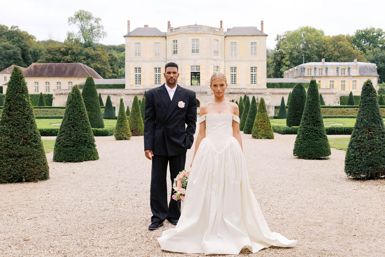 Elegant wedding couple poses in front of a grand French at Chateau de Villette with manicured topiary gardens on a sunny day.
