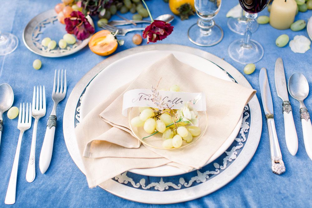 Elegant table setting with blue and white decor featuring wine glasses, silverware, and scattered grapes on a blue tablecloth.