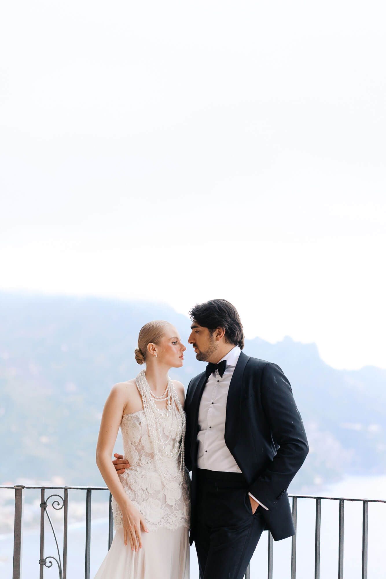 Elegant couple in wedding attire share an intimate moment on a balcony overlooking misty mountains.