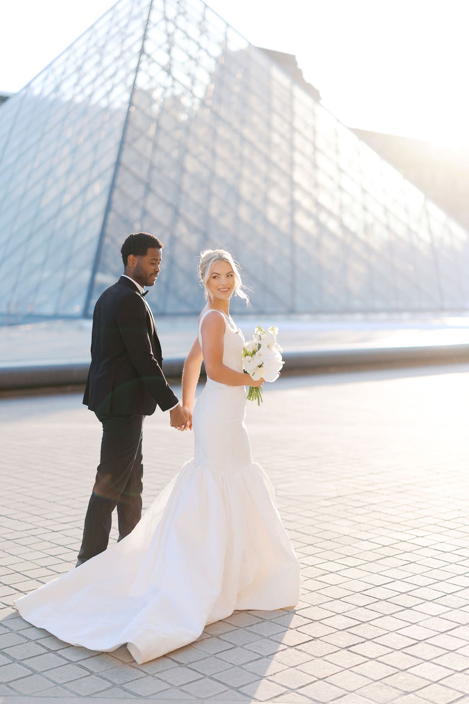 Elegant wedding couple poses together on patterned stone tiles near a modern architectural structure at sunset.