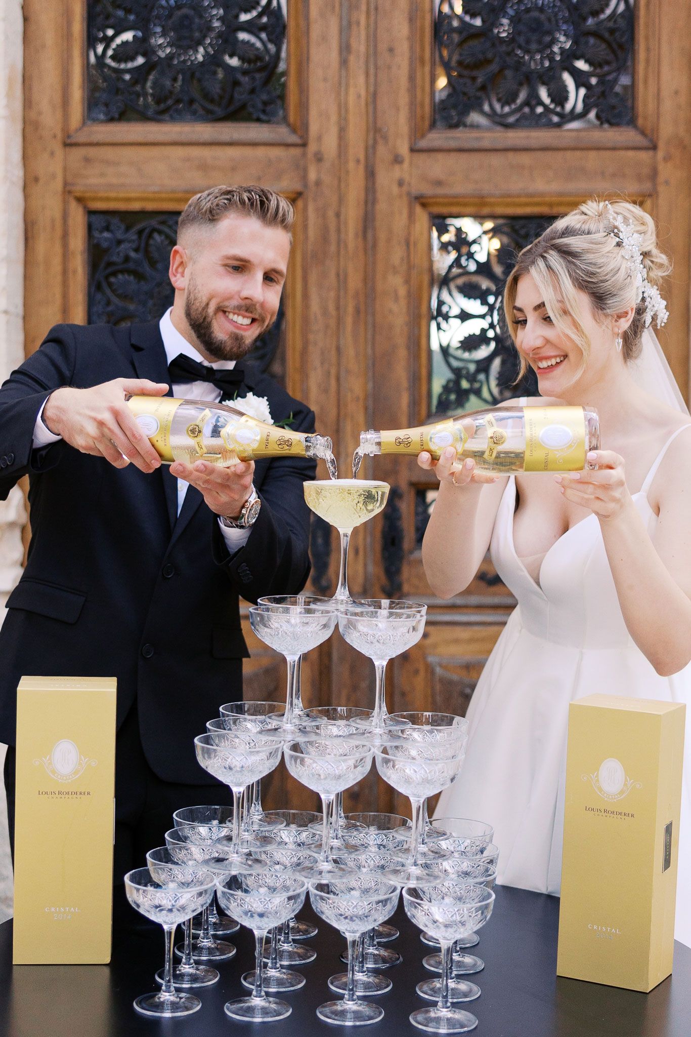 Wedding couple pouring champagne into a tower of stacked coupe glasses during their celebration at Chateau Marinay, France.