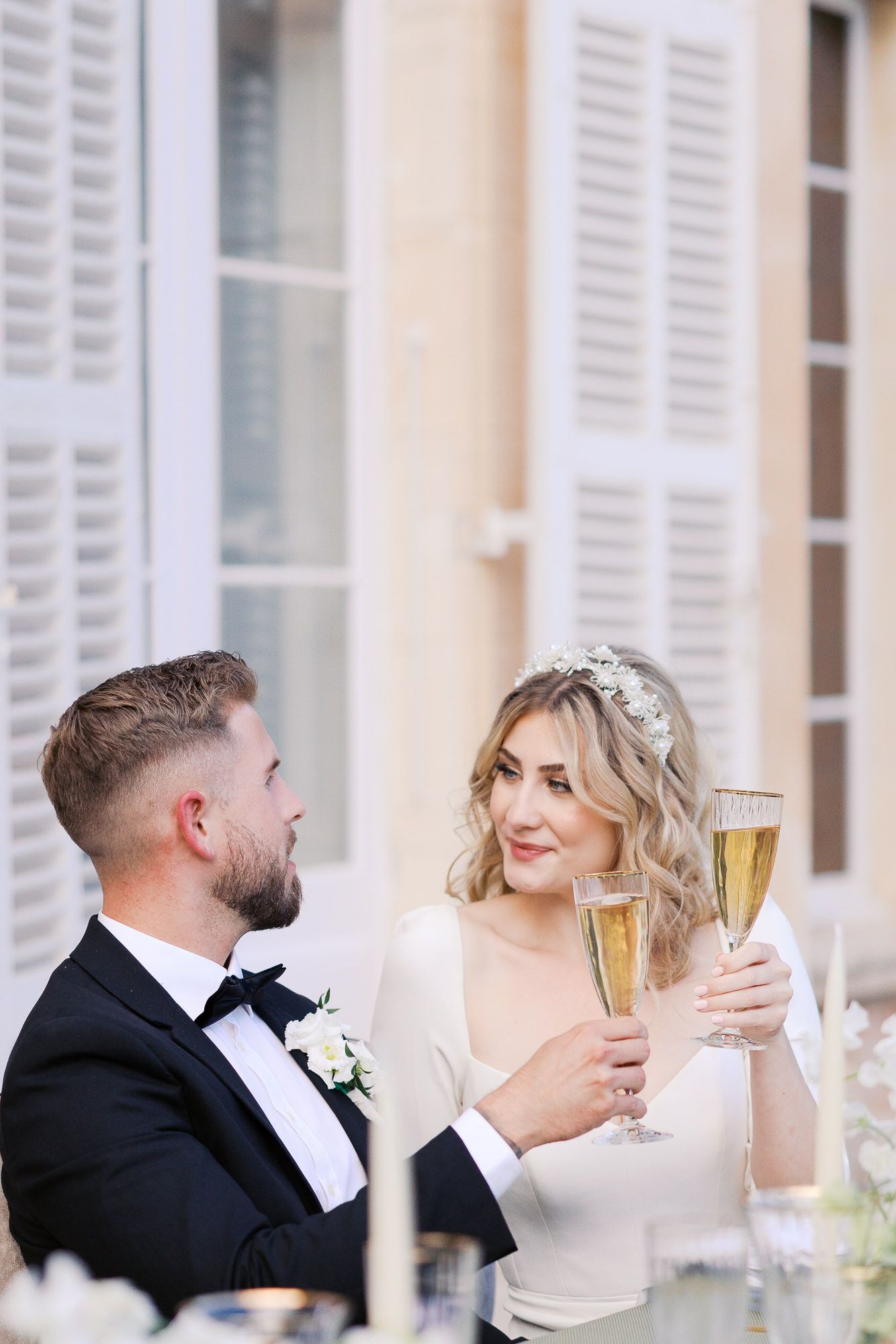 A romantic wedding couple toasts with champagne glasses in front of elegant white shutters at Château Martinay.