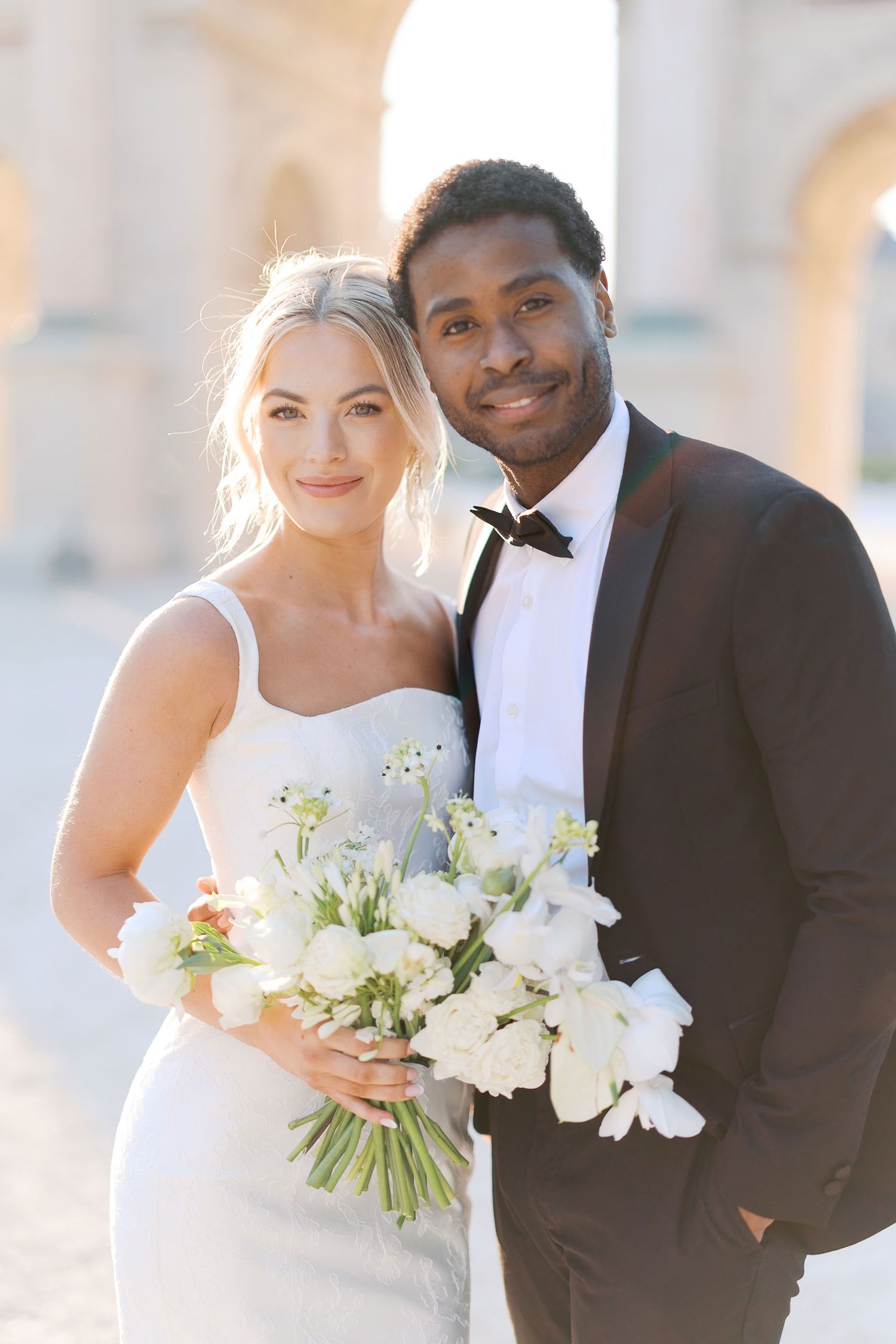 A bride in white dress holds elegant white floral bouquet while standing with groom in black tuxedo outdoors.