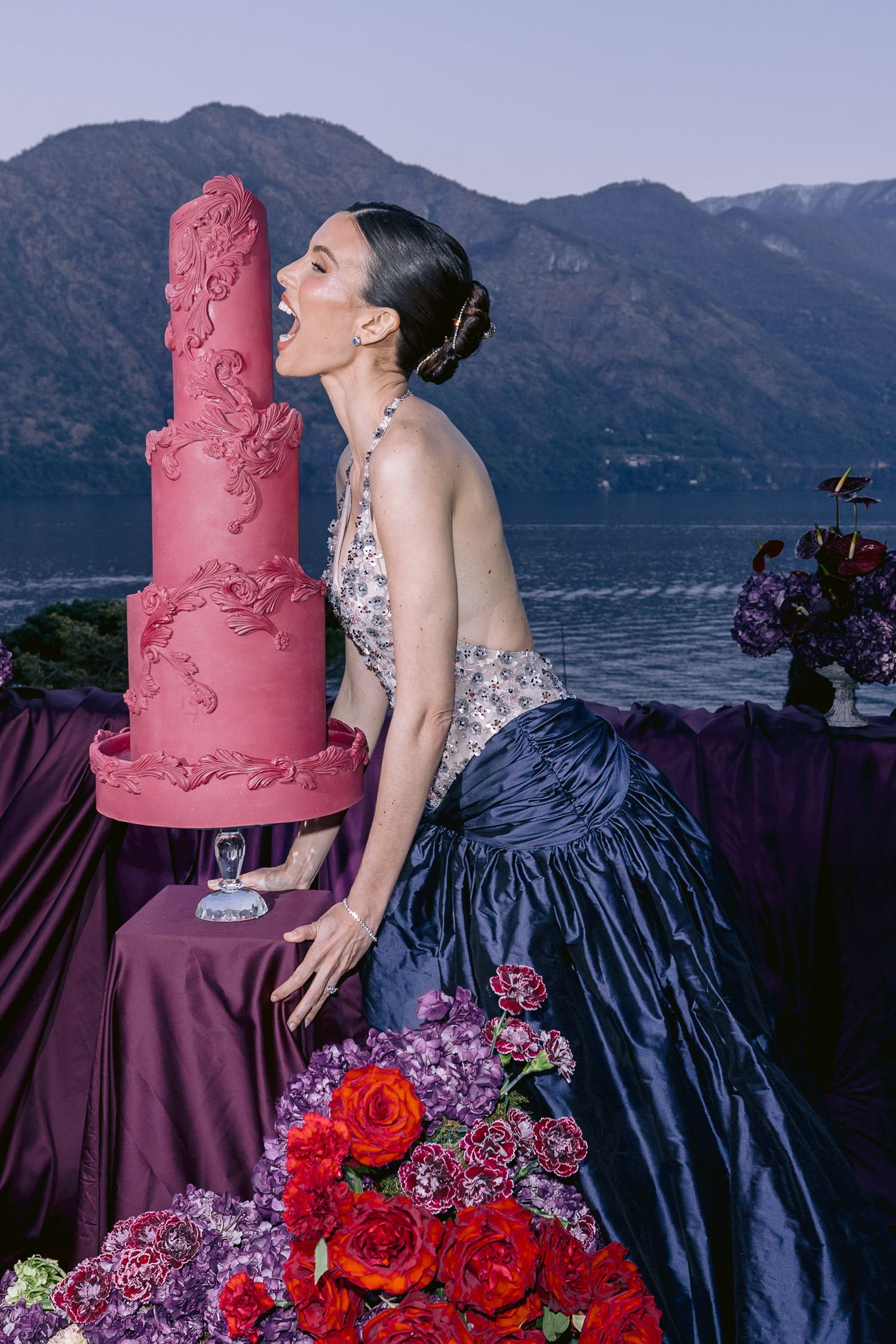 Woman in elegant gown licking a tall pink fondant cake outdoors with mountain lake backdrop.