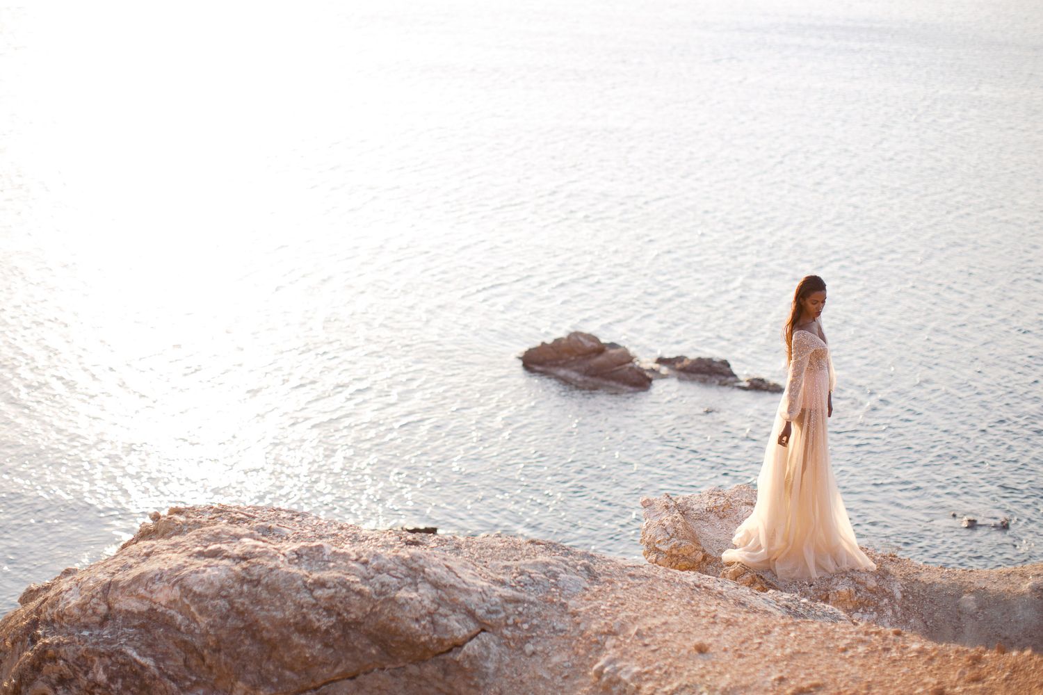 A bride in a flowing white dress stands on a coastal cliff edge overlooking the serene Mediterranean Sea at sunset.