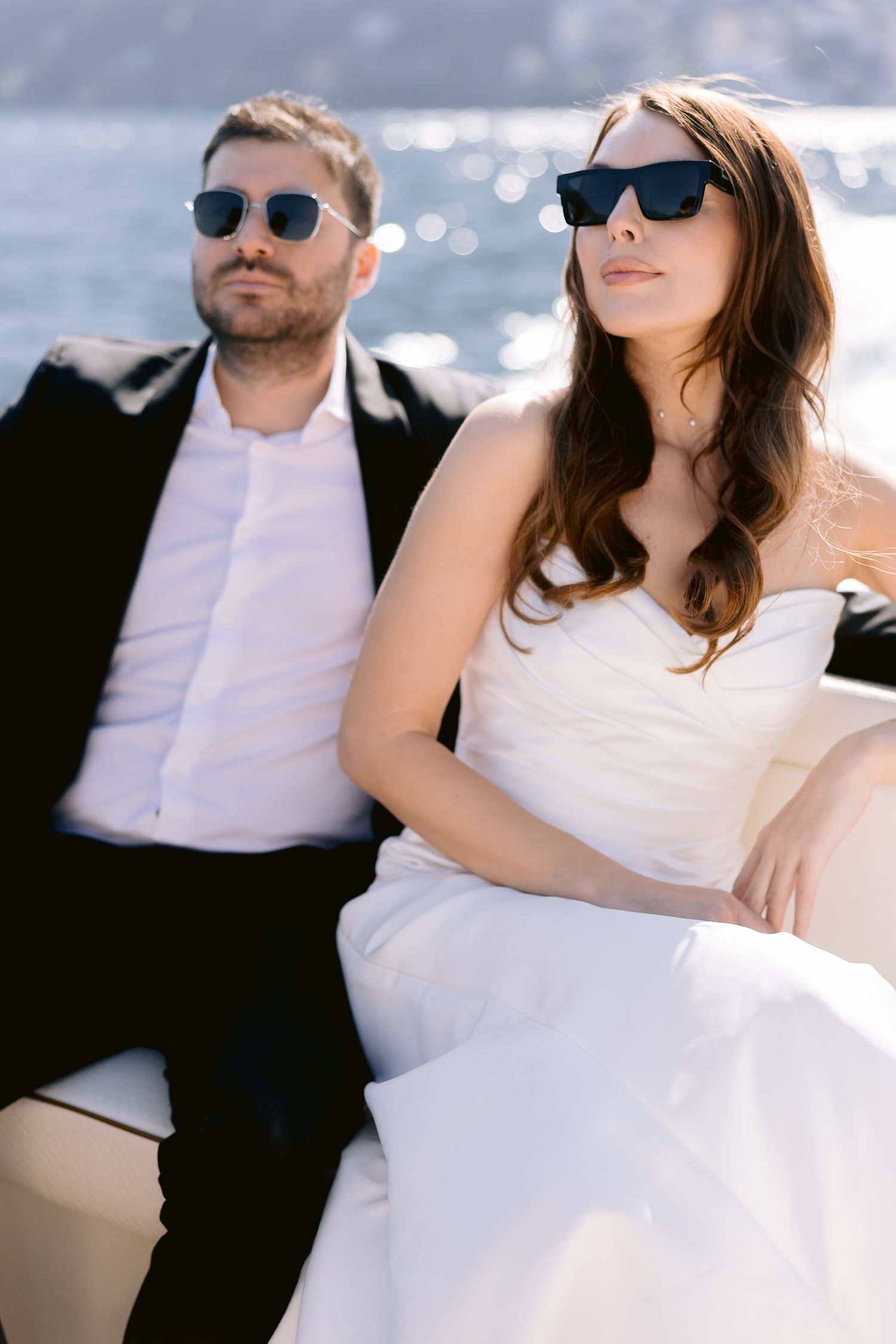 Bride and groom in sunglasses sitting on a yacht, wearing wedding attire with sparkling water in the background.