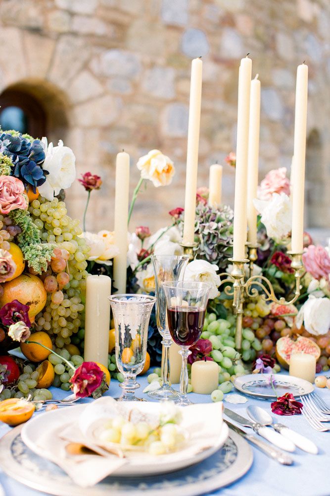 Romantic table setting with cream candles, grapes, and pink flowers against stone wall.