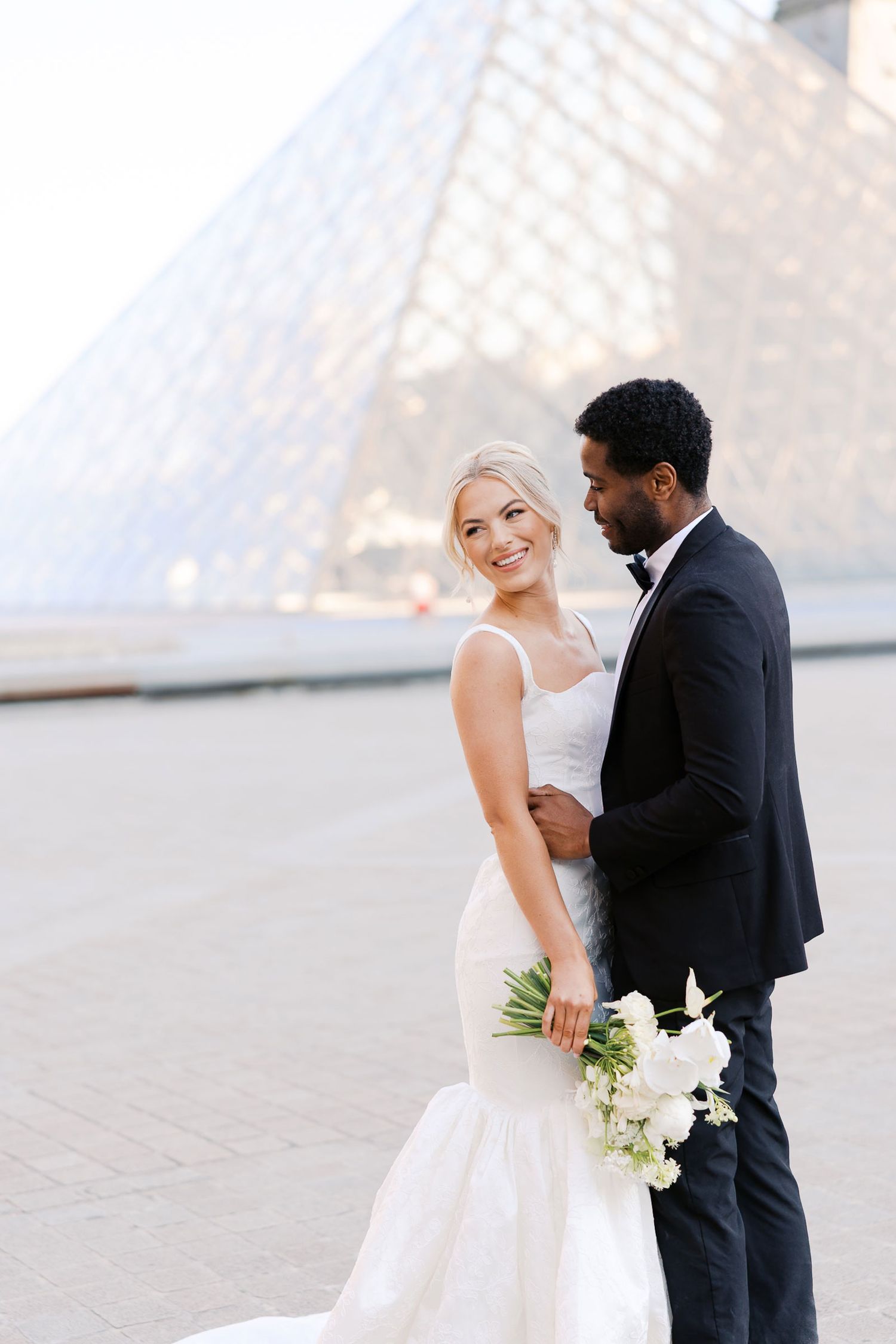 A romantic wedding couple shares intimate moments in front of the Louvre pyramid in Paris.