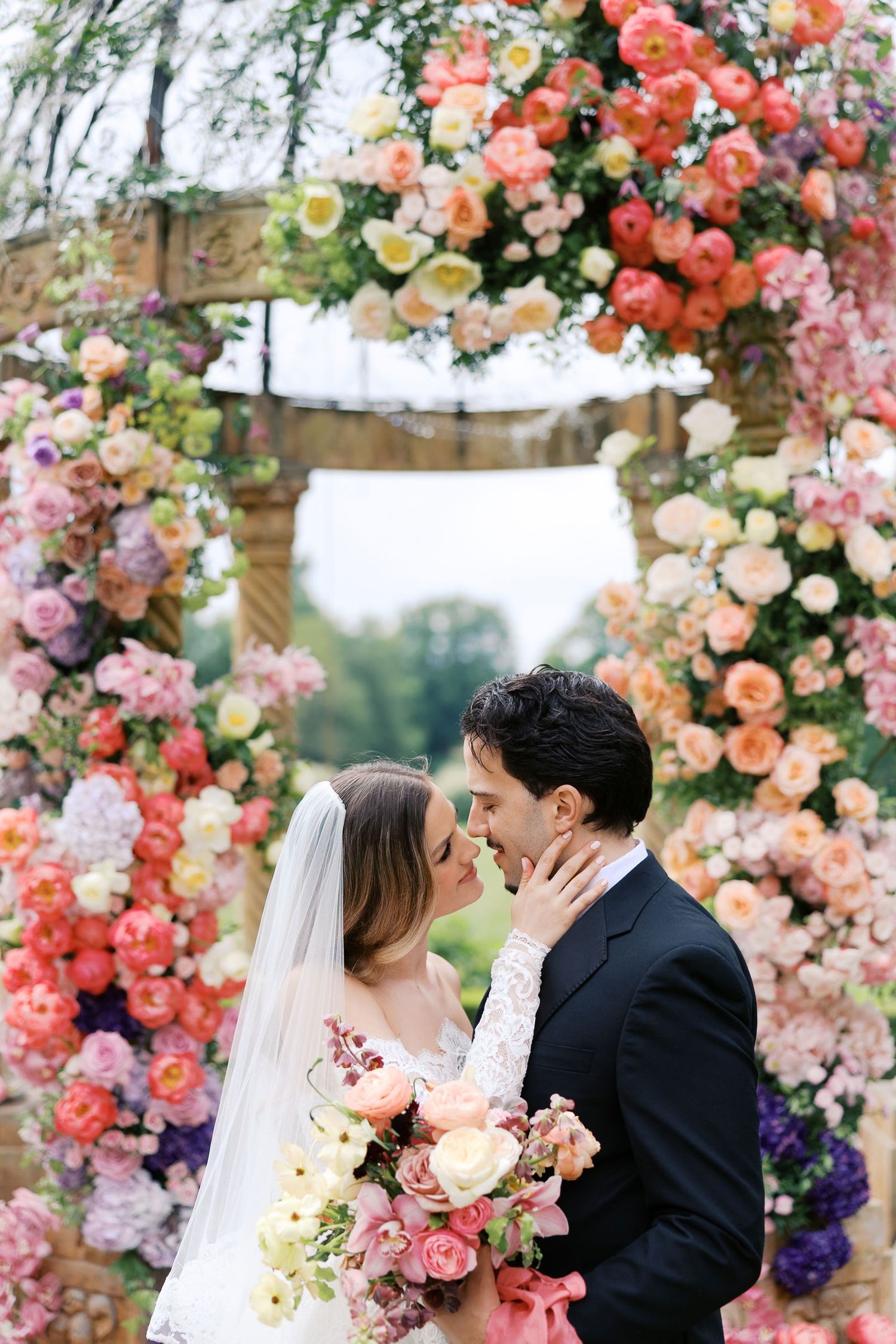 A romantic wedding couple shares a kiss under a stunning floral arch adorned with coral, pink and purple blooms at Chateau de Challain.