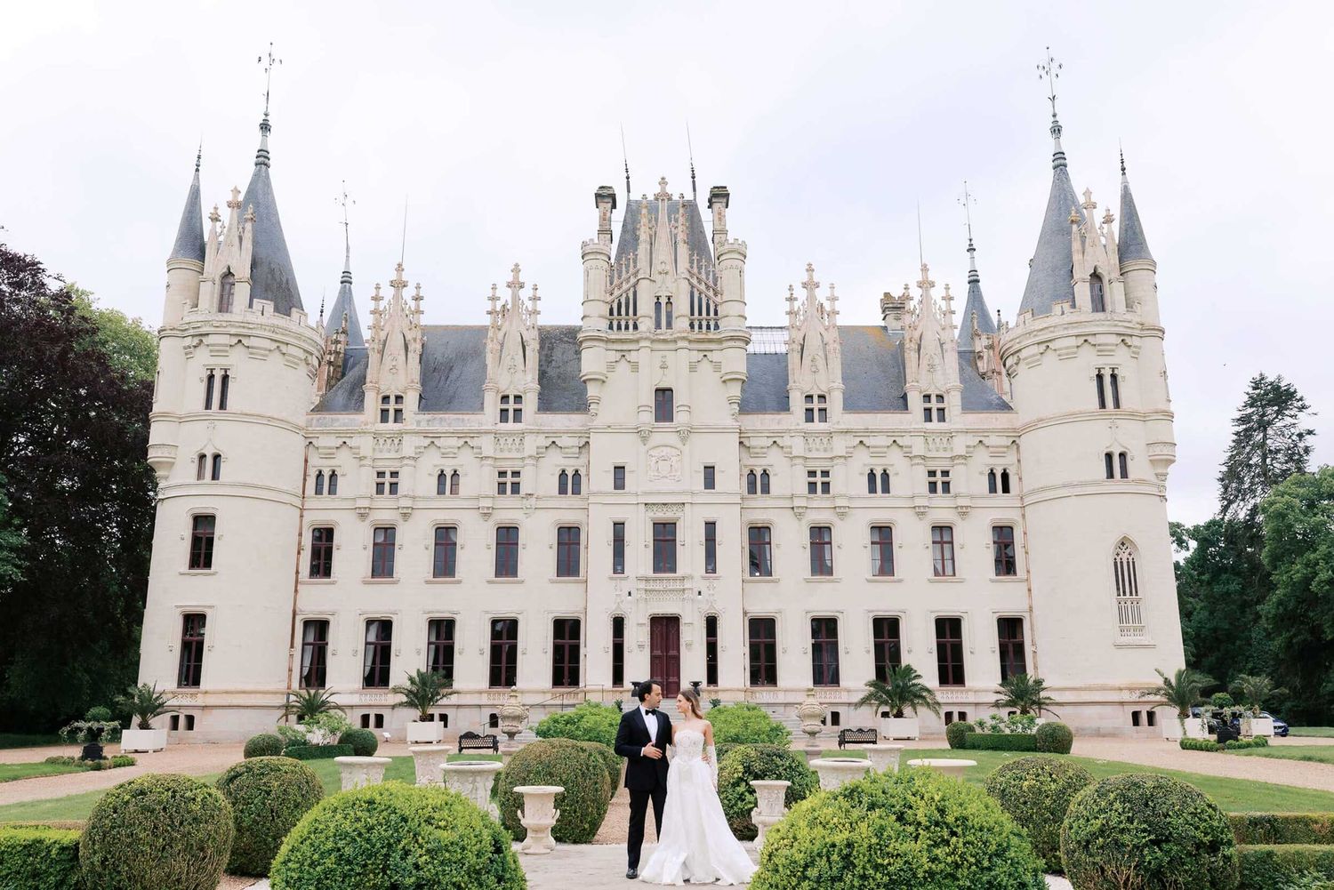 A wedding couple stands before the grand white Chateau Challain castle in France with its Gothic spires and manicured gardens.