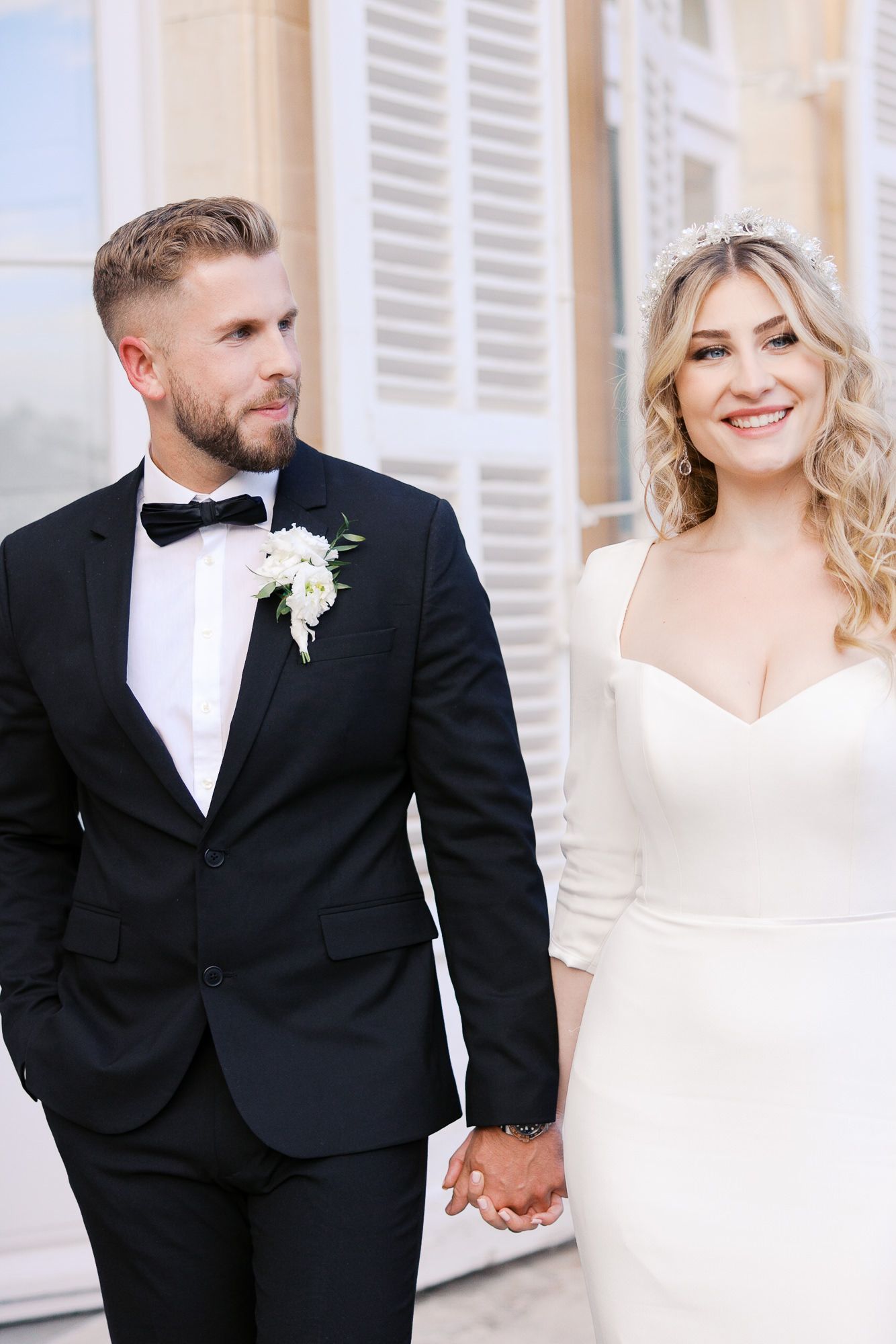 A bride in a sleek white dress and groom in black tuxedo hold hands while walking together outdoors at Château Martinay.