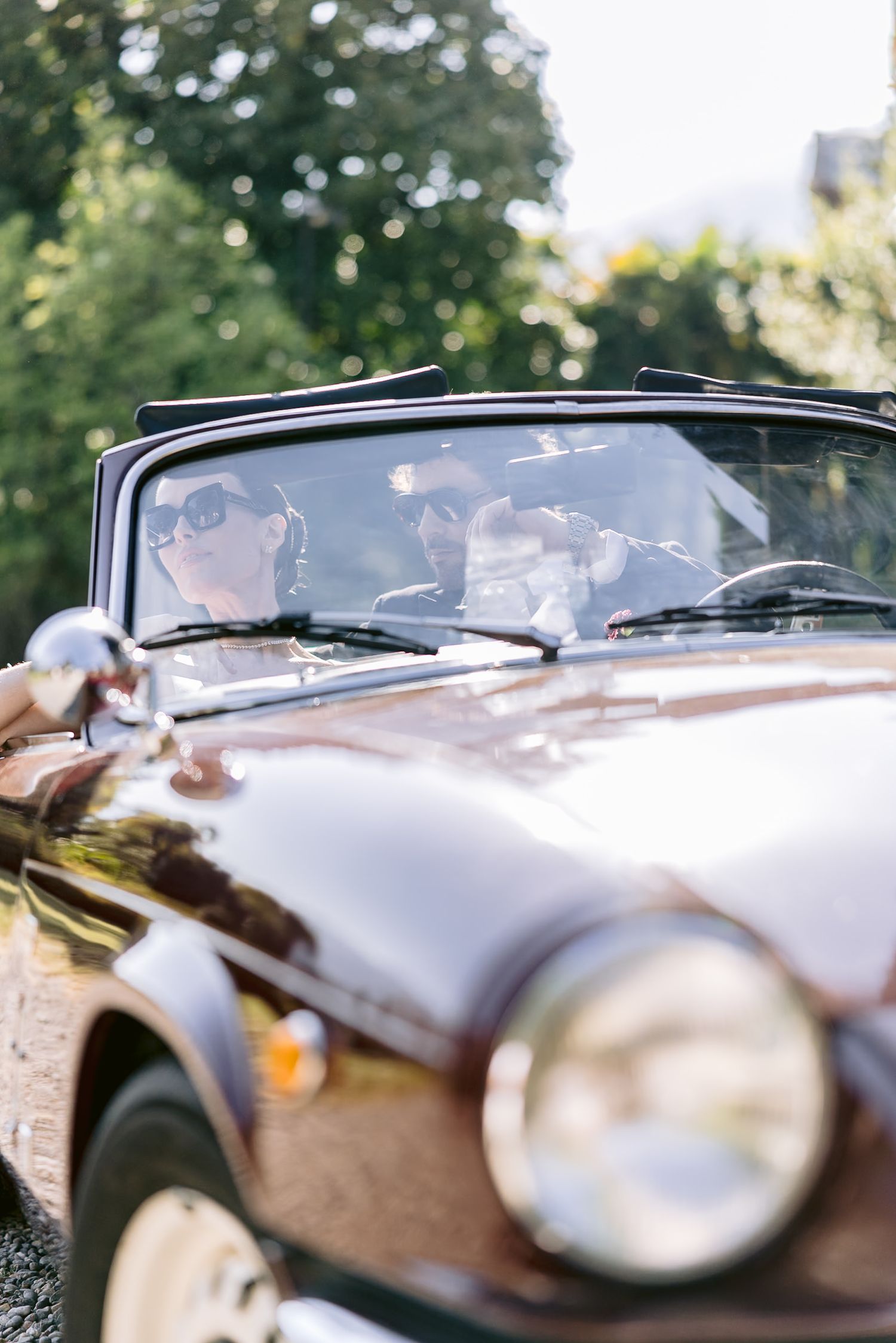 Couple riding in a vintage convertible car on a sunny day, woman wearing sunglasses in the passenger seat.