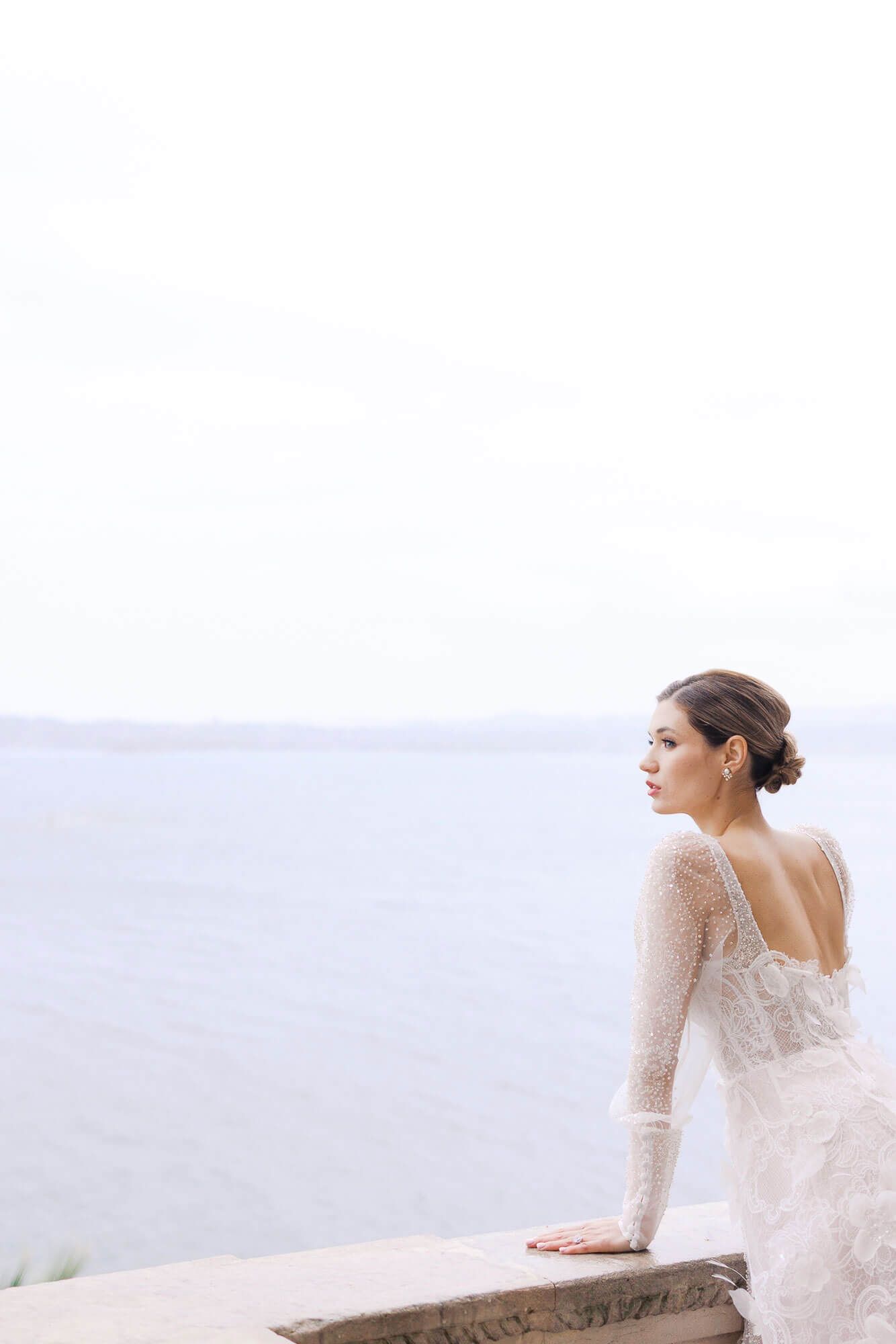 A bride in a long-sleeved lace wedding dress gazes into the distance while sitting on a stone wall with misty views at Isola del Garda.
