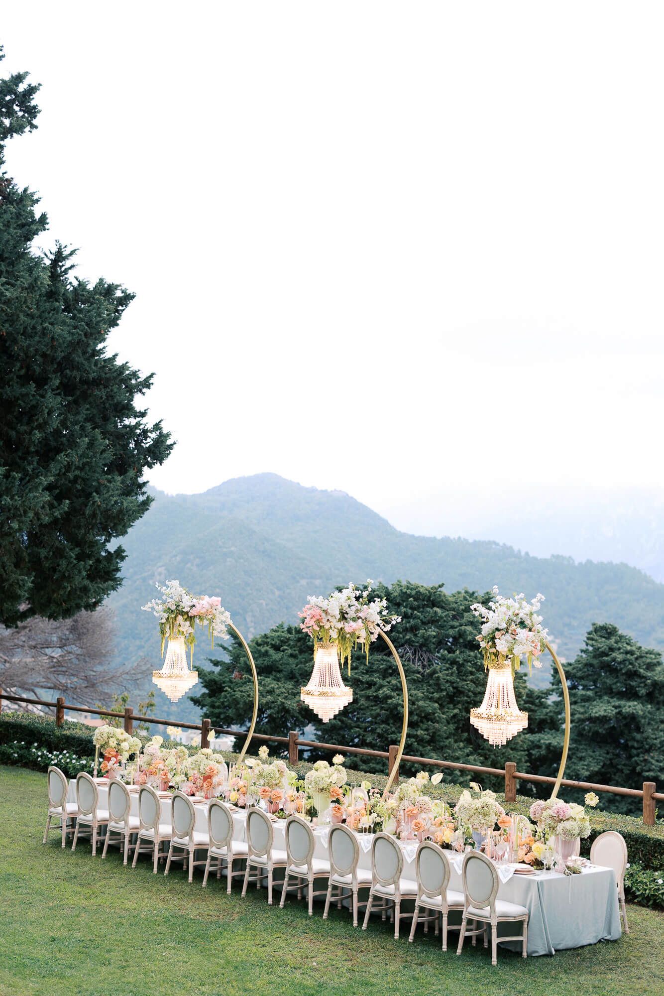 Elegant outdoor wedding reception setup with long white tables and floral arrangements against a scenic mountain backdrop.