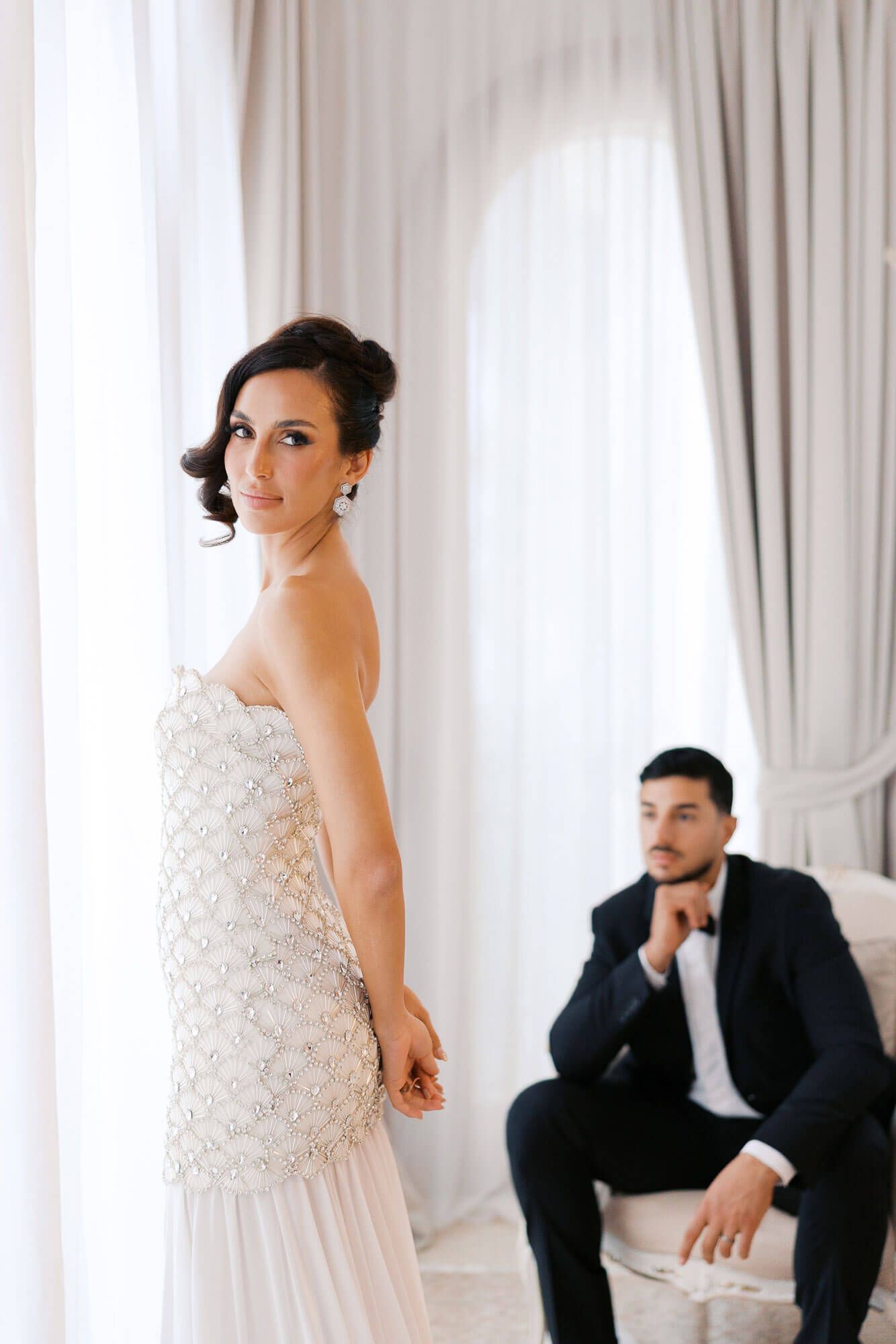 Elegant bride in white lace gown poses thoughtfully while groom in dark suit sits in bright modern room.