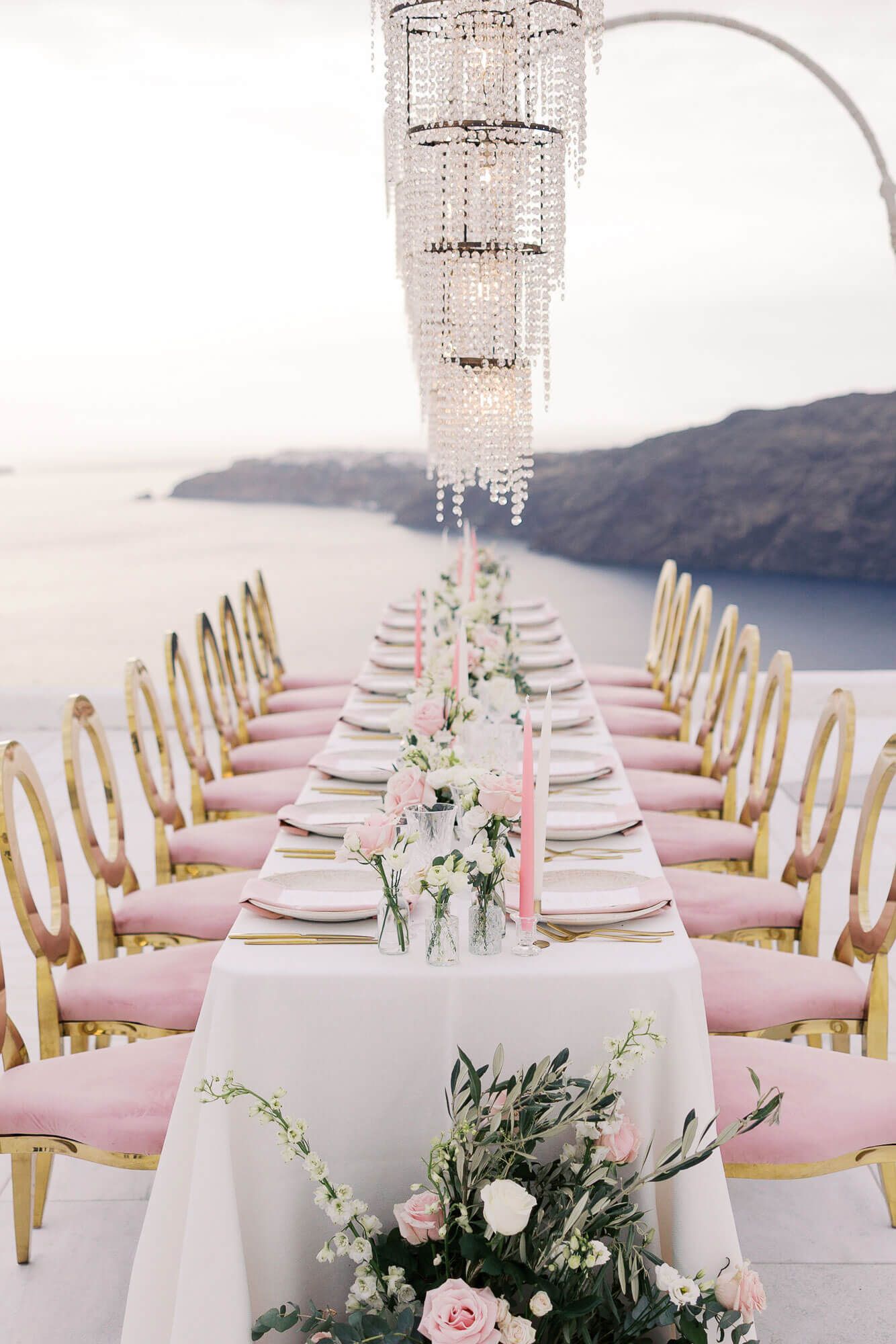 Elegant outdoor wedding reception setup with white tables, gold chairs, and pink floral centerpieces overlooking Mediterranean sunset views.
