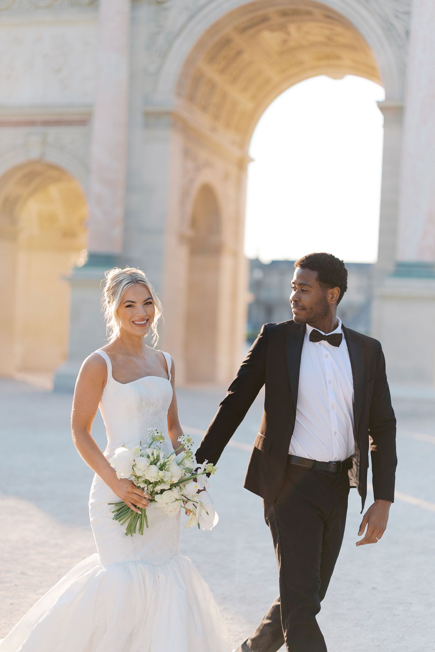 A bride in white dress and groom in tuxedo walk through an elegant arched hallway in romantic wedding photos.