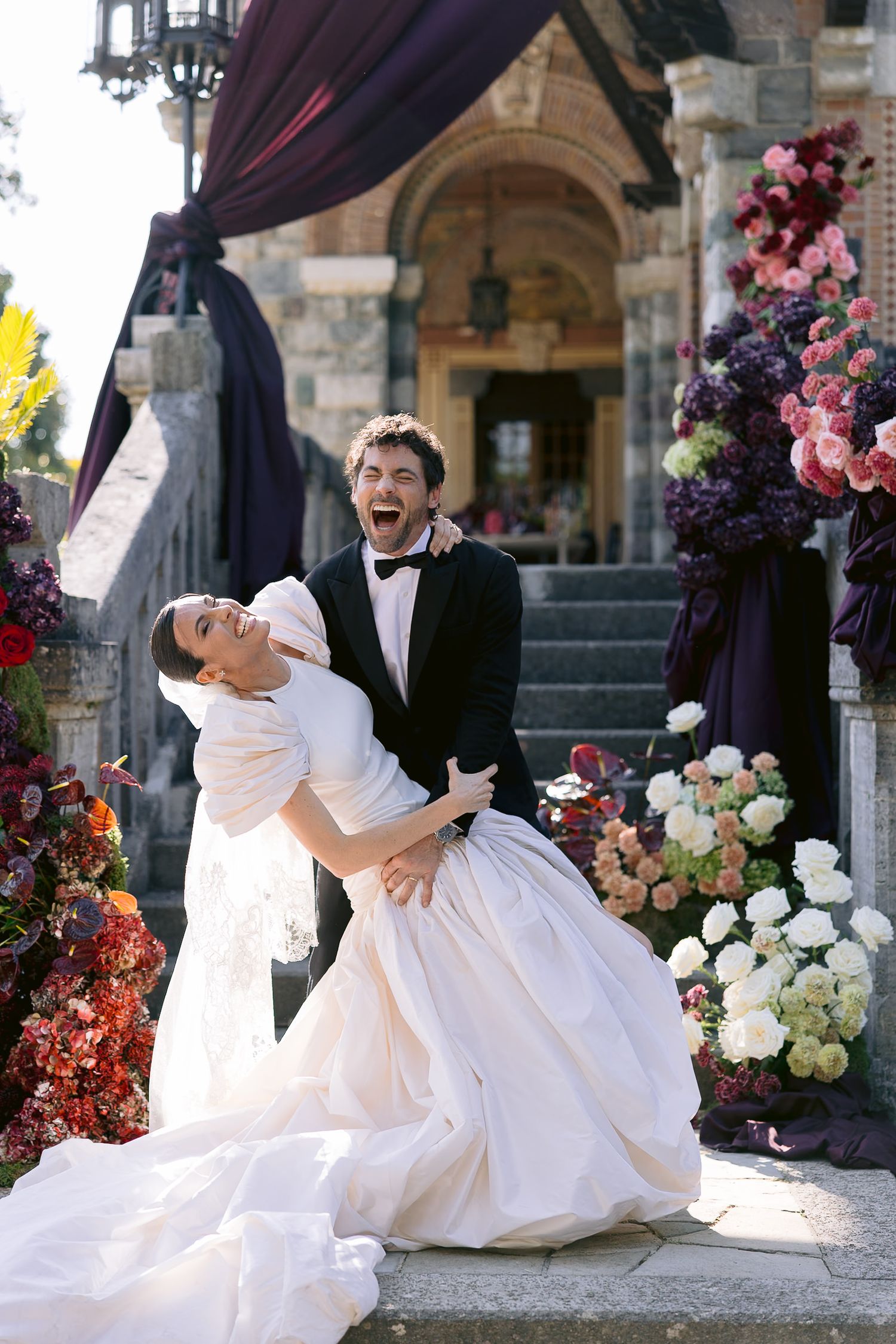 Joyful bride and groom laughing outdoors at floral-decorated wedding venue with stone building backdrop.