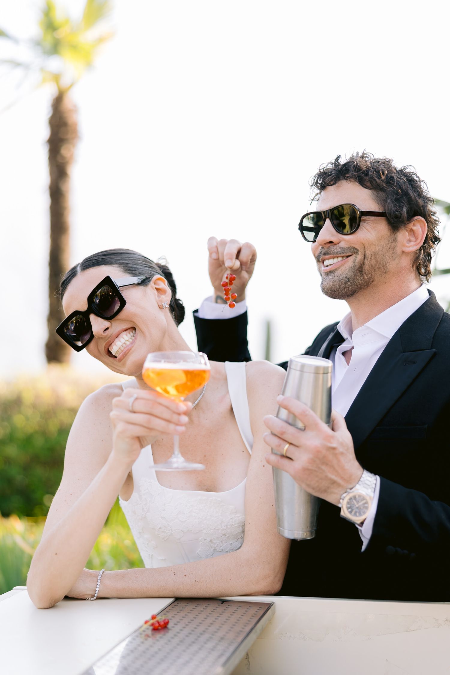 Happy couple in sunglasses celebrating outdoors, bride holding aperol spritz and groom with cocktail shaker near palm trees.