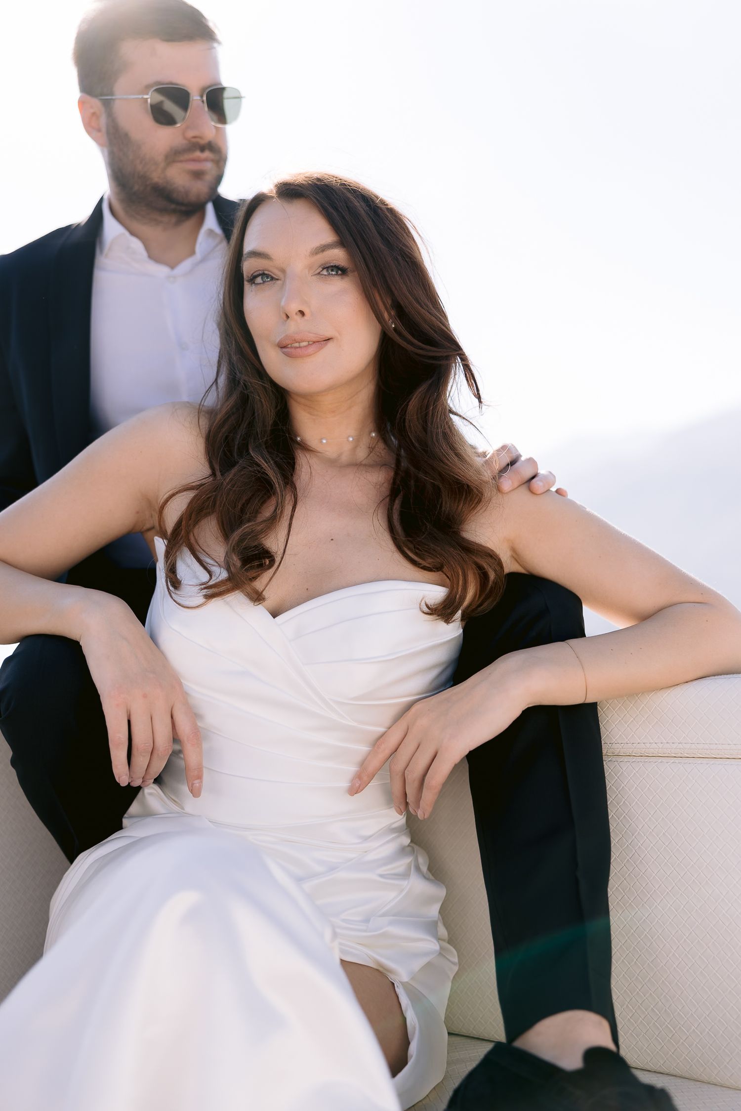 Elegant bride in strapless white gown sits with groom in dark suit and sunglasses during outdoor wedding photo session.