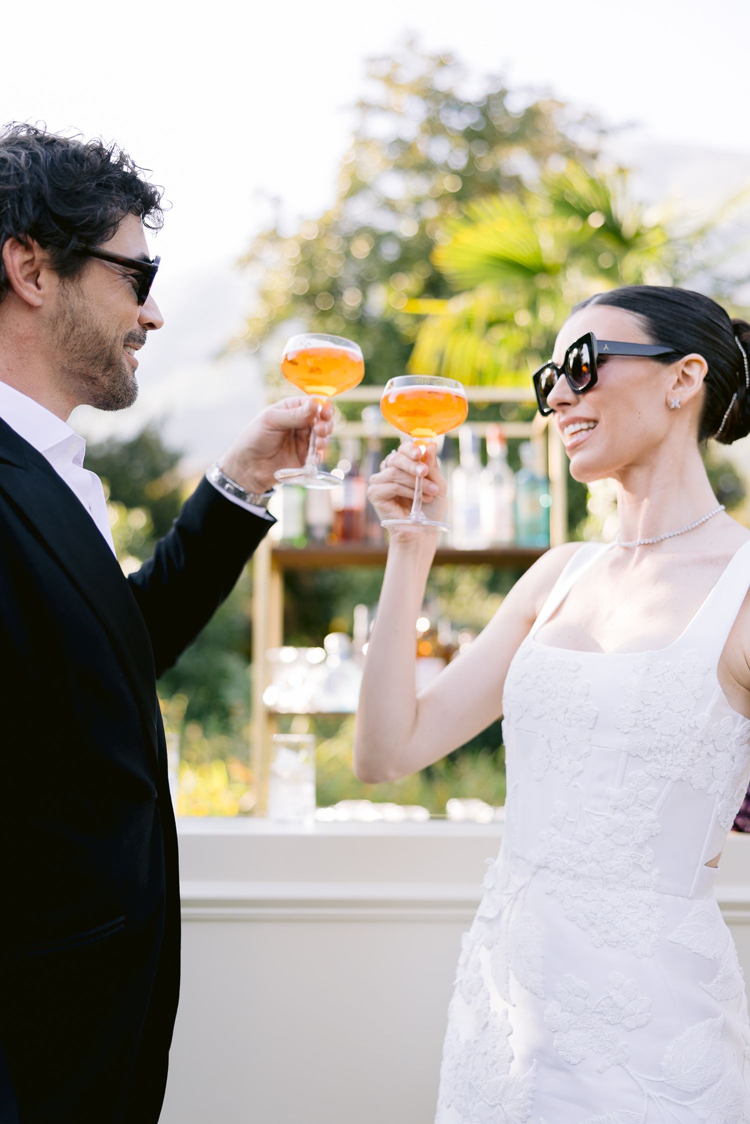Bride and groom in sunglasses toasting with orange cocktails at outdoor wedding reception.