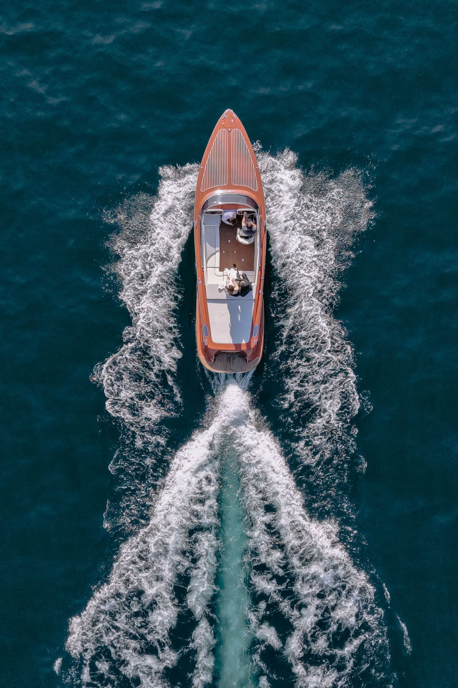 Aerial view of a classic wooden speedboat cutting through deep blue water, leaving a white wake trail.