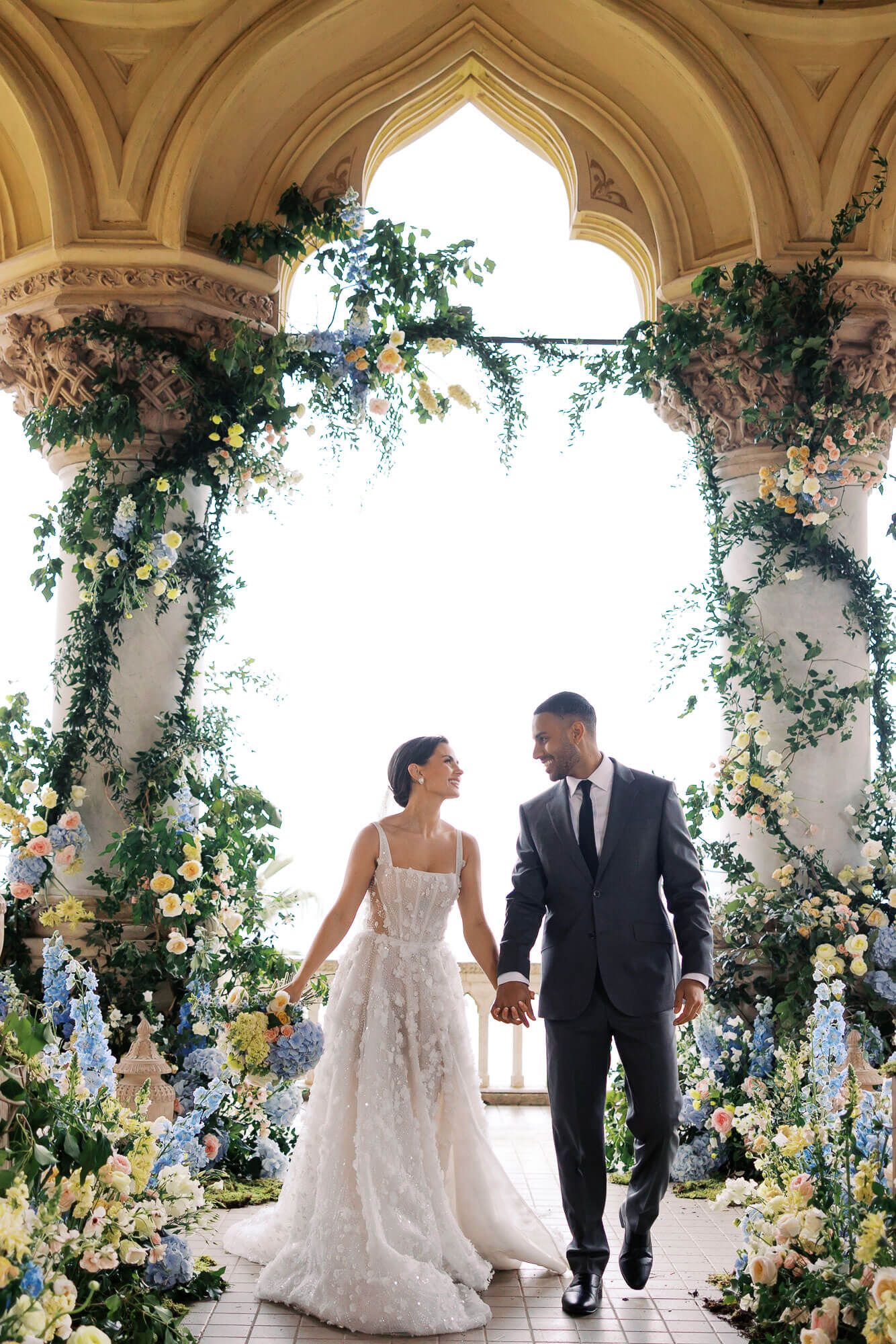 Elegant wedding couple walking through ornate archway decorated with cascading blue and white floral arrangements at Isola del Garda.