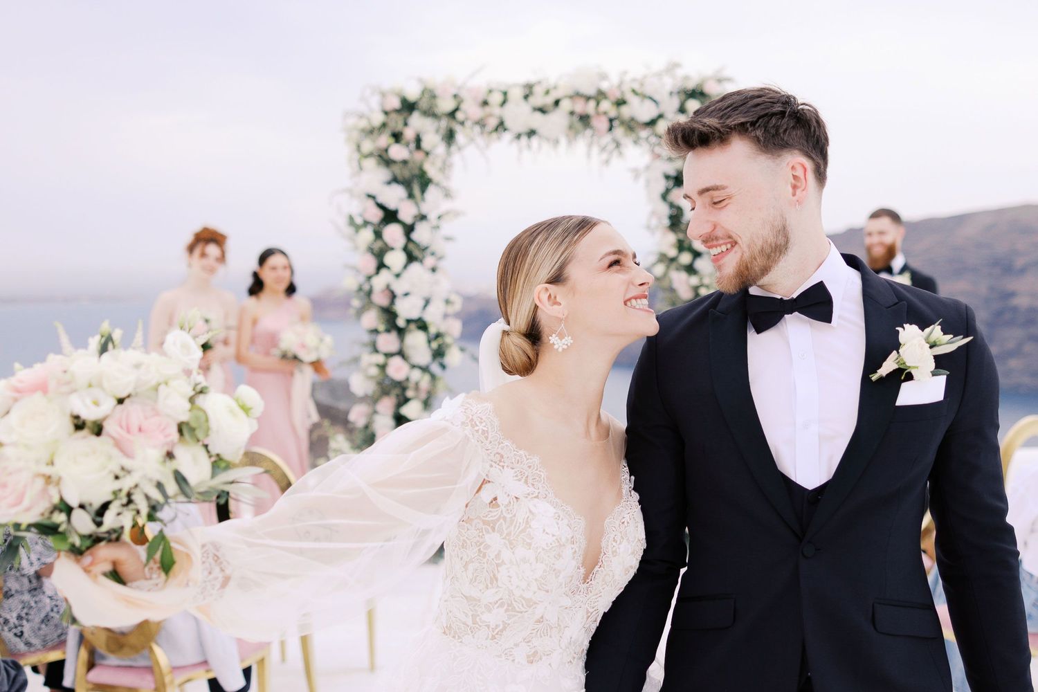 Romantic wedding couple shares intimate moments in black and white formal attire surrounded by floral arrangements in Rocabella Santorini.