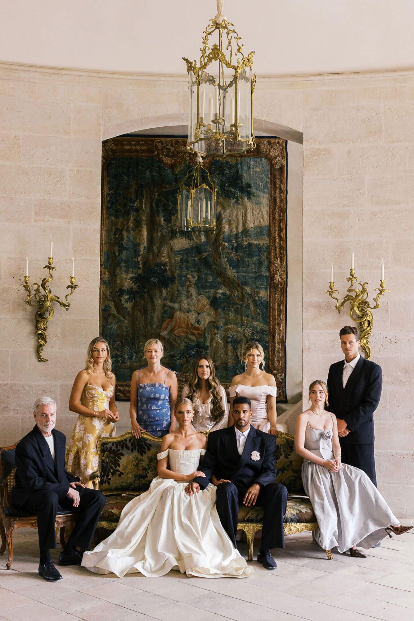 An elegant bridal party seated on a bench in a light-filled room with chandeliers at Chateau de Villette.