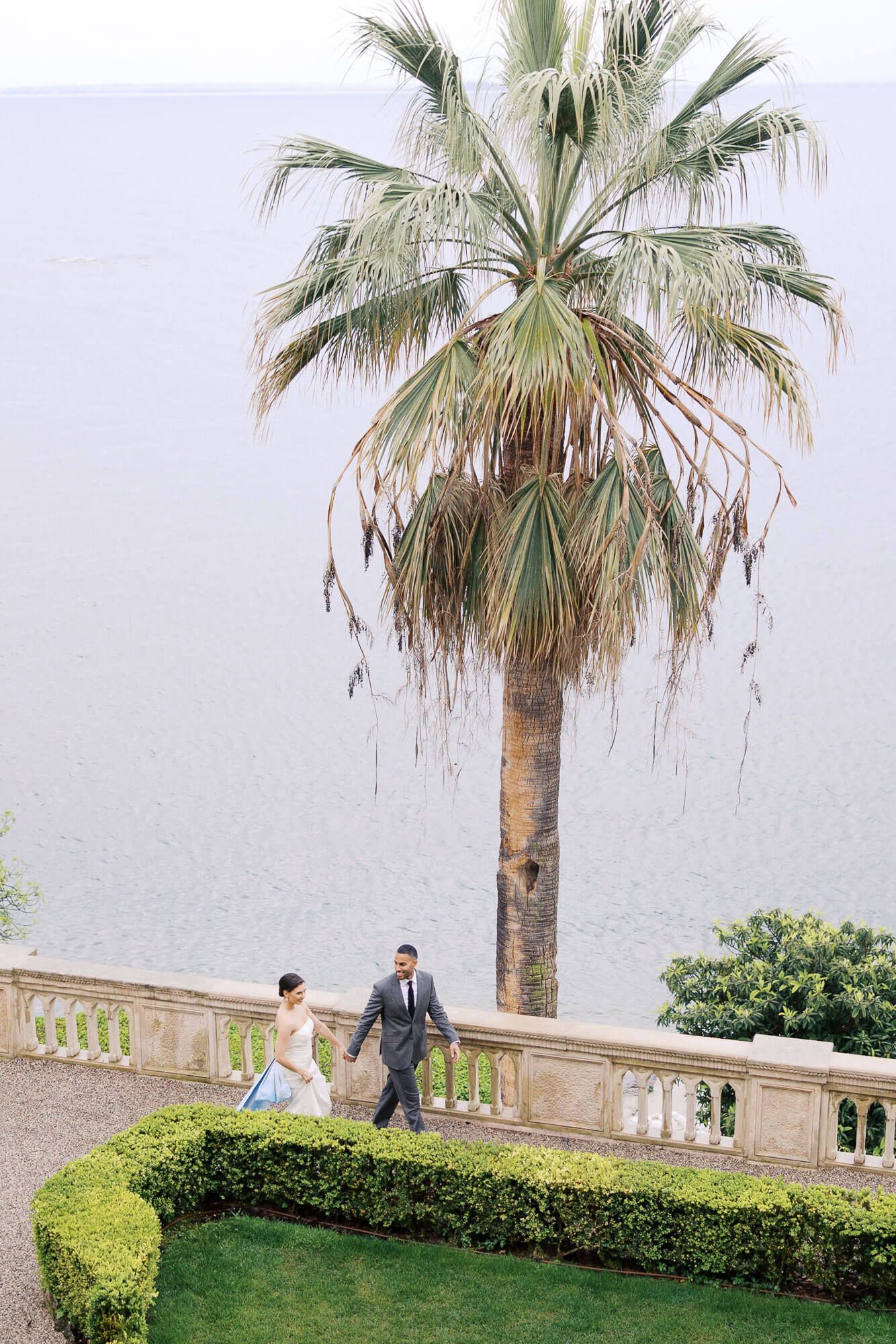 Two people stroll along a scenic Mediterranean terrace with palm trees and stone balustrades overlooking the sea at Isola del Garda.