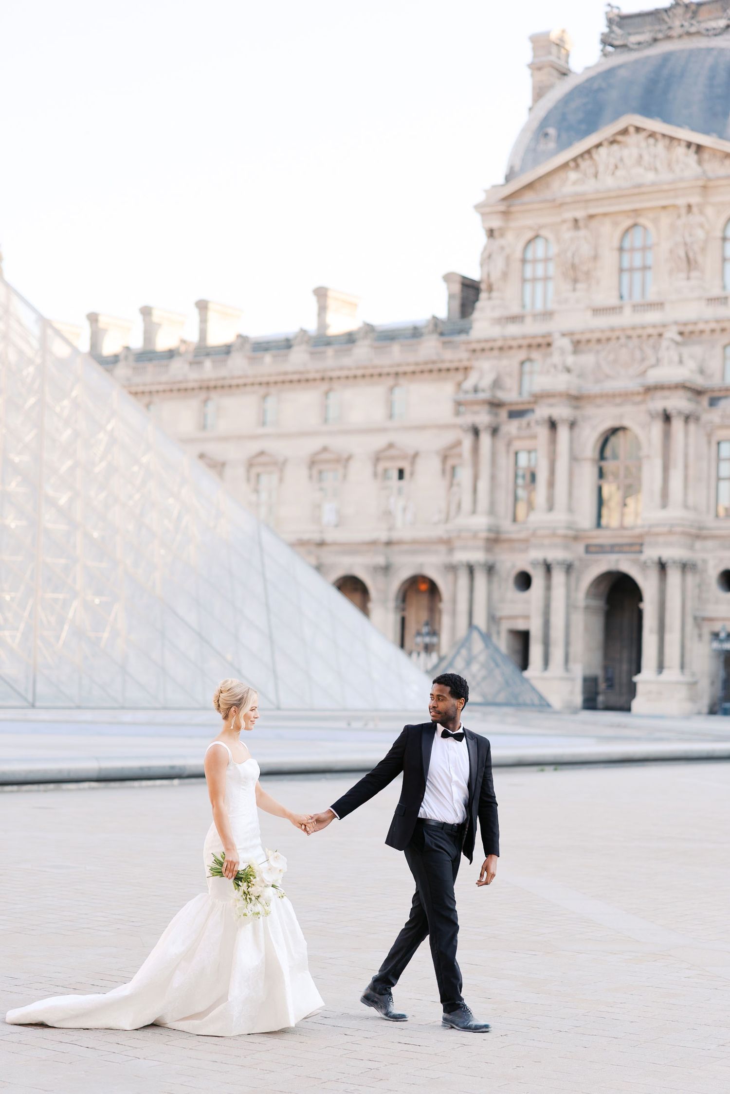 A wedding couple in wedding attire playfully poses in front of the iconic Louvre Pyramid in Paris on a bright sunny day.