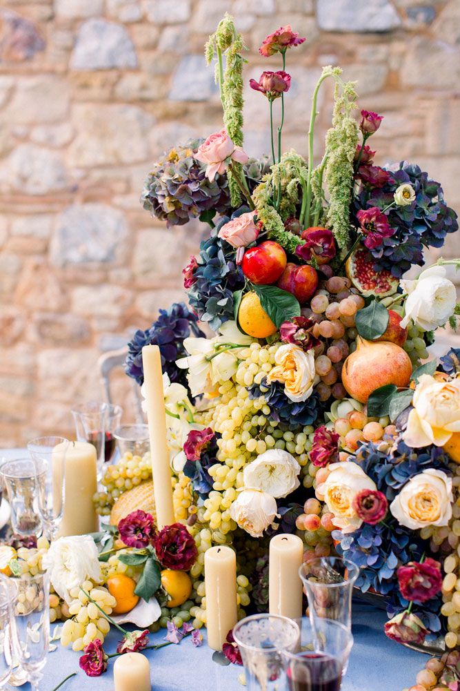 Elegant table setting with lush floral centerpiece featuring roses, grapes, citrus fruits and navy blue hydrangeas with white candles.