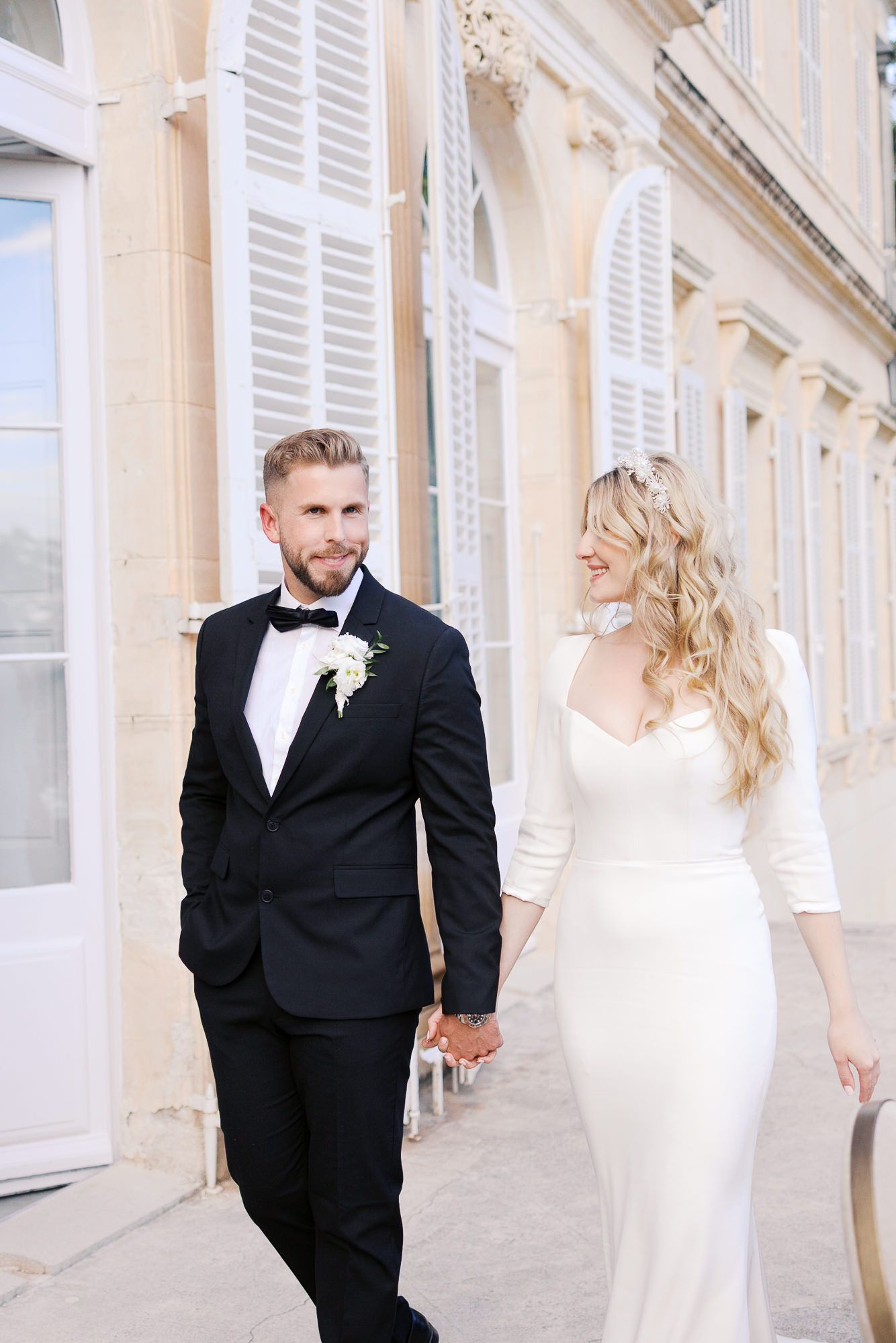 Elegant couple in wedding attire walking together outside a French-style building with white shutters at Château Martinay.