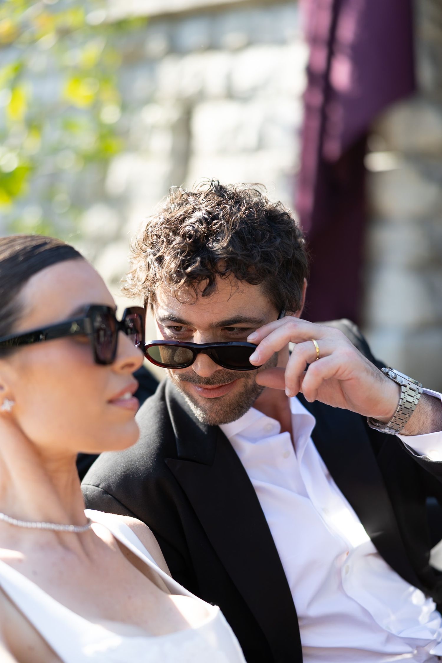 Elegant couple in sunglasses outdoors, man in black suit adjusting shades, woman in white dress with pearl necklace.